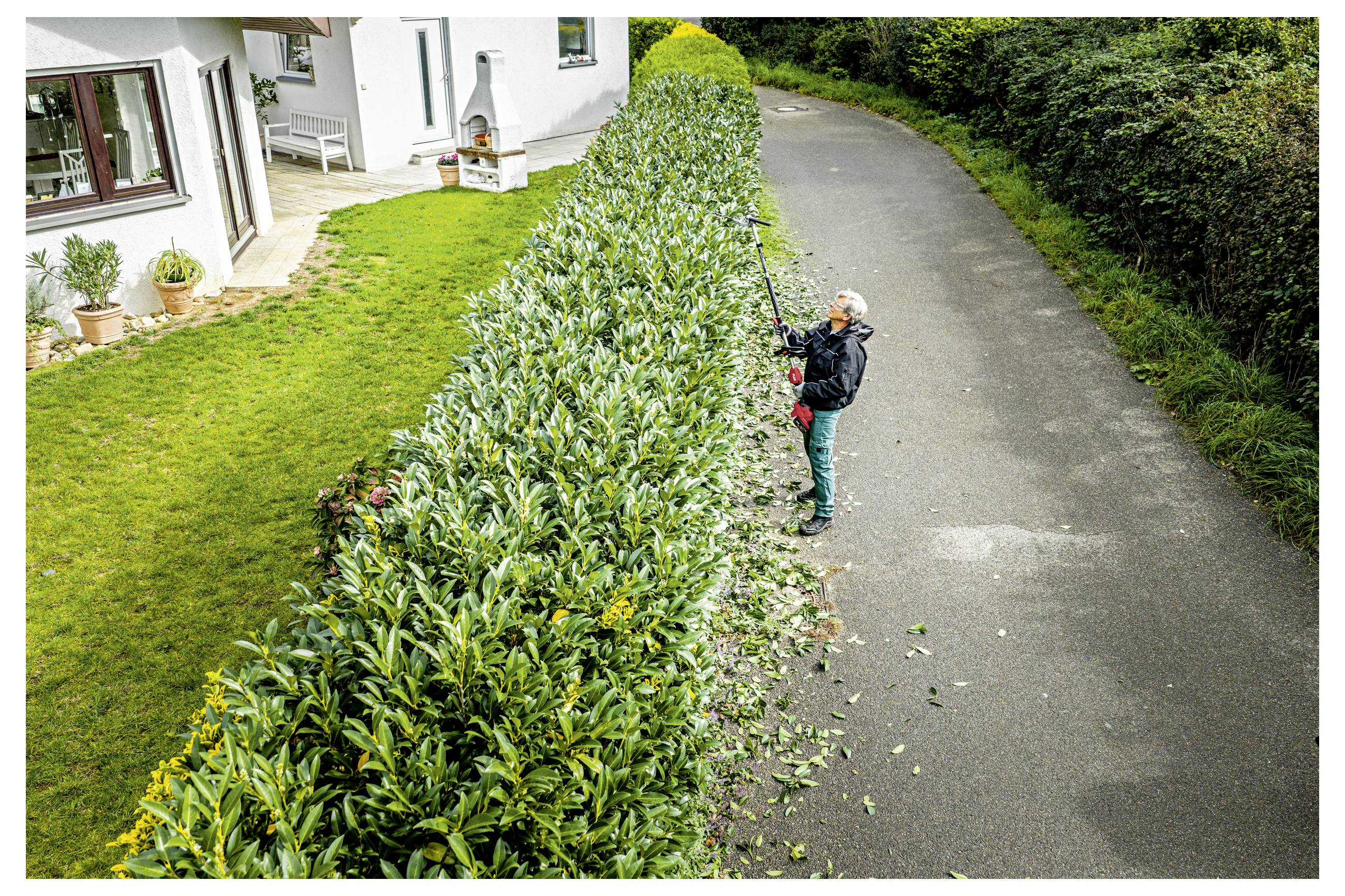 Eine Person schneidet eine hohe Hecke entlang einer Einfahrt neben einem weißen Haus, mit Schnittgut auf dem Gehweg.