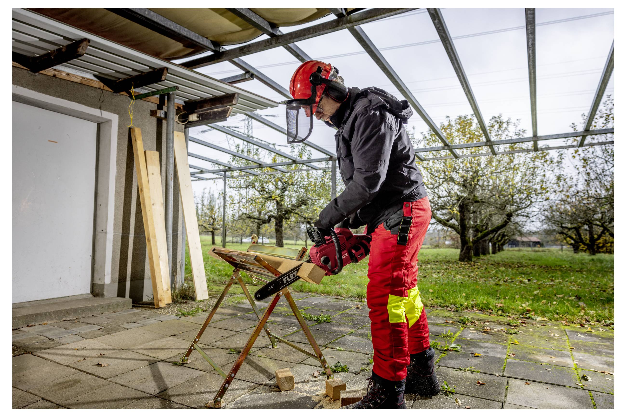 Eine Person in Schutzausrüstung schneidet Holz auf einem Sägebock mit einer Kettensäge unter einem überdachten Außenarbeitsbereich, mit Gartenbäumen im Hintergrund.