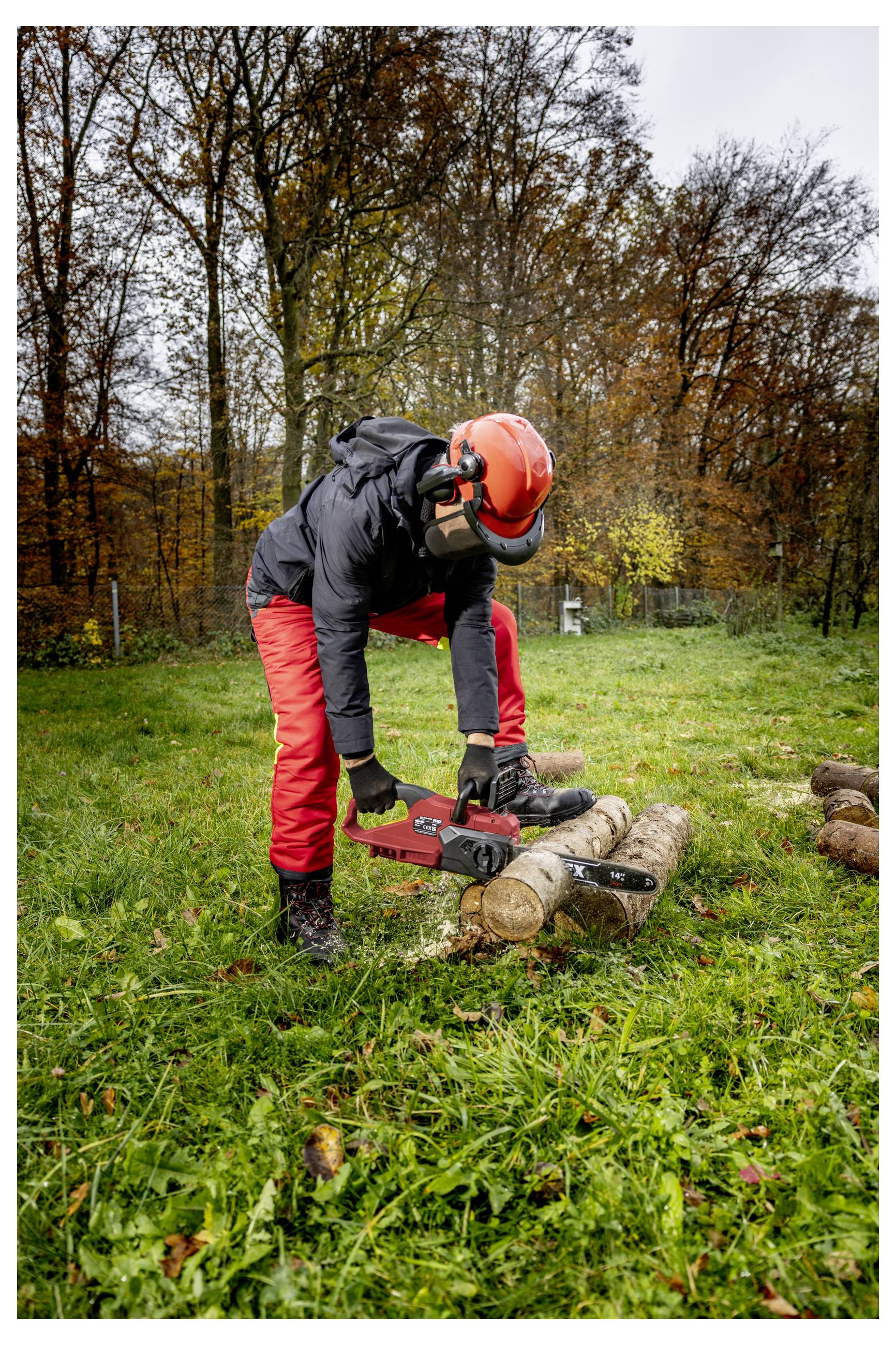 Eine Person in Schutzausrüstung nutzt eine Kettensäge, um Holzstämme auf Gras in einem bewaldeten Gebiet zu zerteilen.