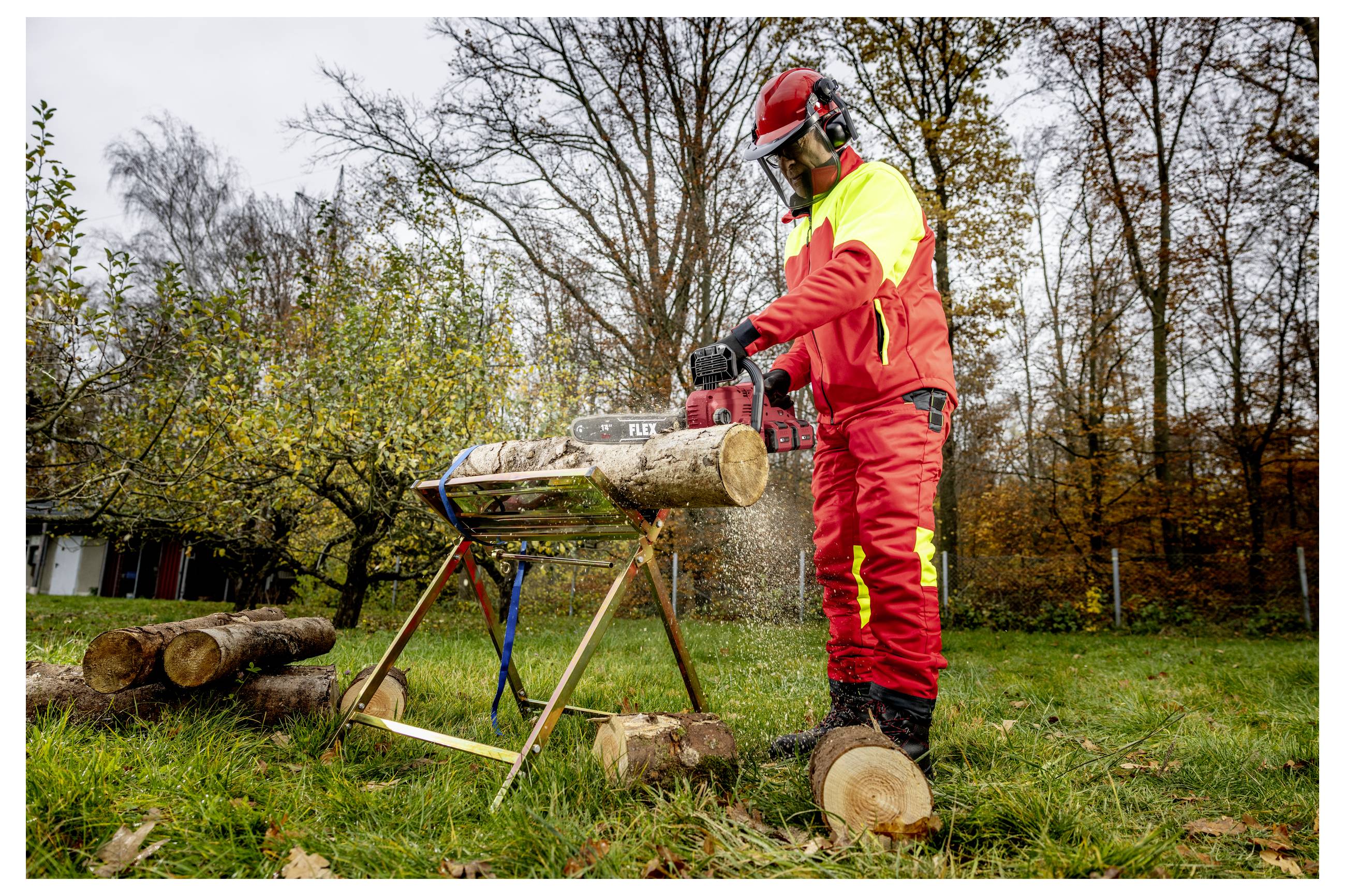 Ein Arbeiter in roter Schutzausrüstung schneidet ein Holzstück mit einer Kettensäge im Freien auf einem grasbewachsenen Gelände, mit gestapelten Holzstämmen und Bäumen im Hintergrund.