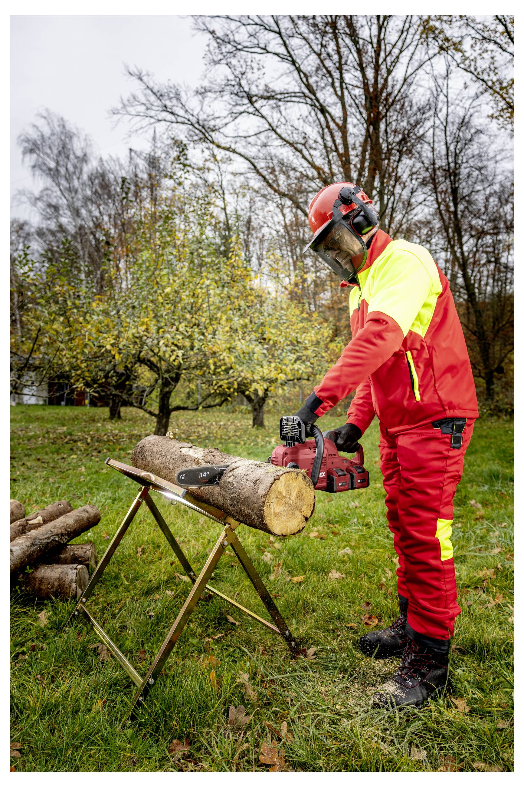Arbeiter in roter Schutzausrüstung, der mit einer Kettensäge einen Baumstamm auf einem Sägebock im Freien auf einer grünen Wiese zuschneidet.