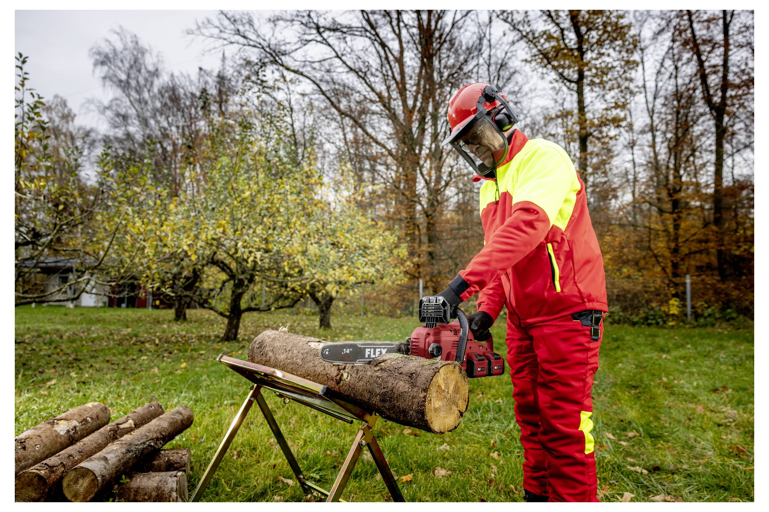Ein Arbeiter in roter Schutzausrüstung schneidet ein Holzstück mit einer Kettensäge auf einem Ständer in einem grasbewachsenen Außenbereich mit gefallenen Ästen in der Nähe.