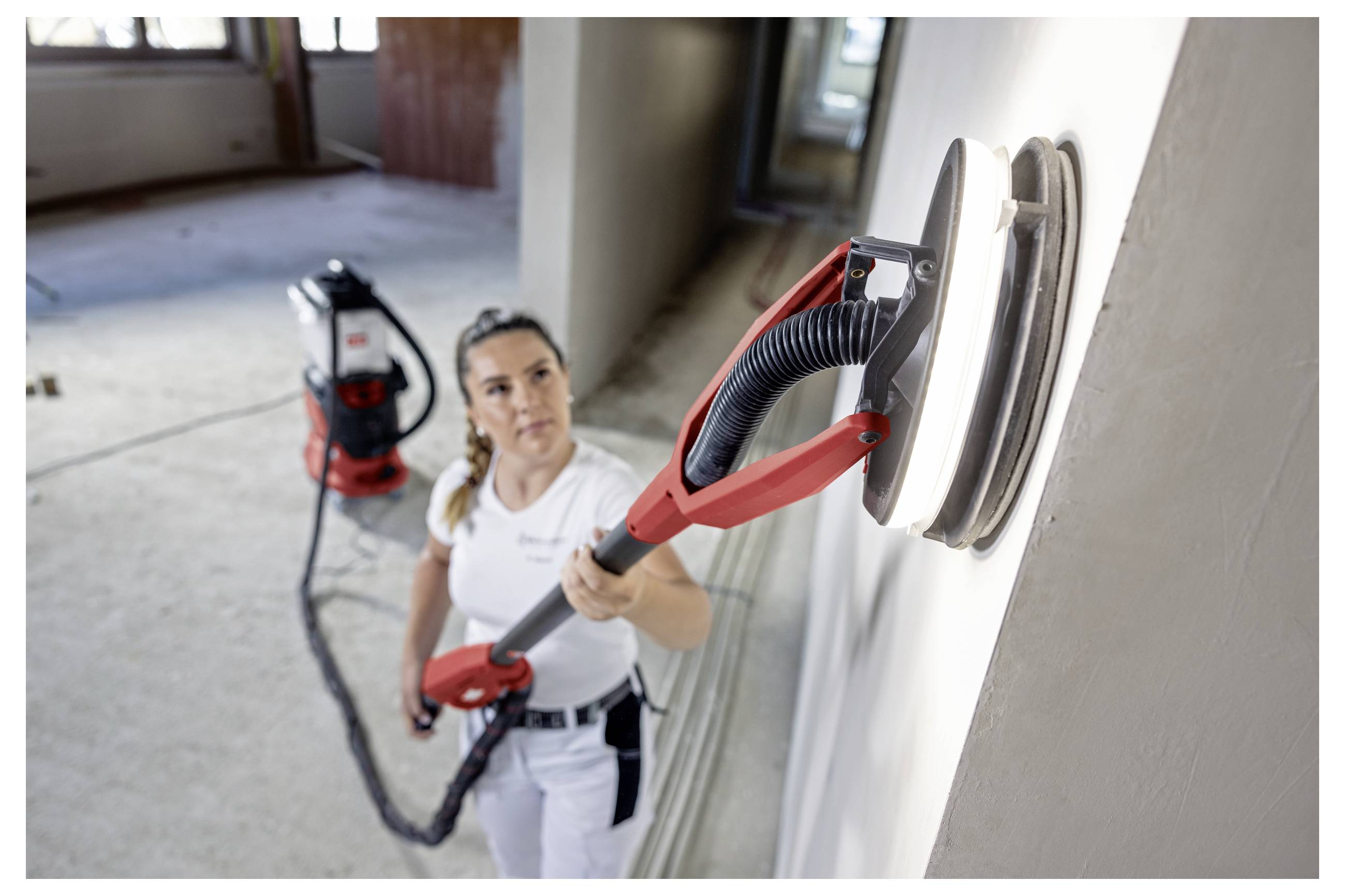 Arbeiter, der einen wandmontierten Trockenbauschleifer verwendet, um eine Innenwand zu glätten, mit angeschlossenem Staubsaugerschlauch und einem Werkstattstaubsauger im Hintergrund.