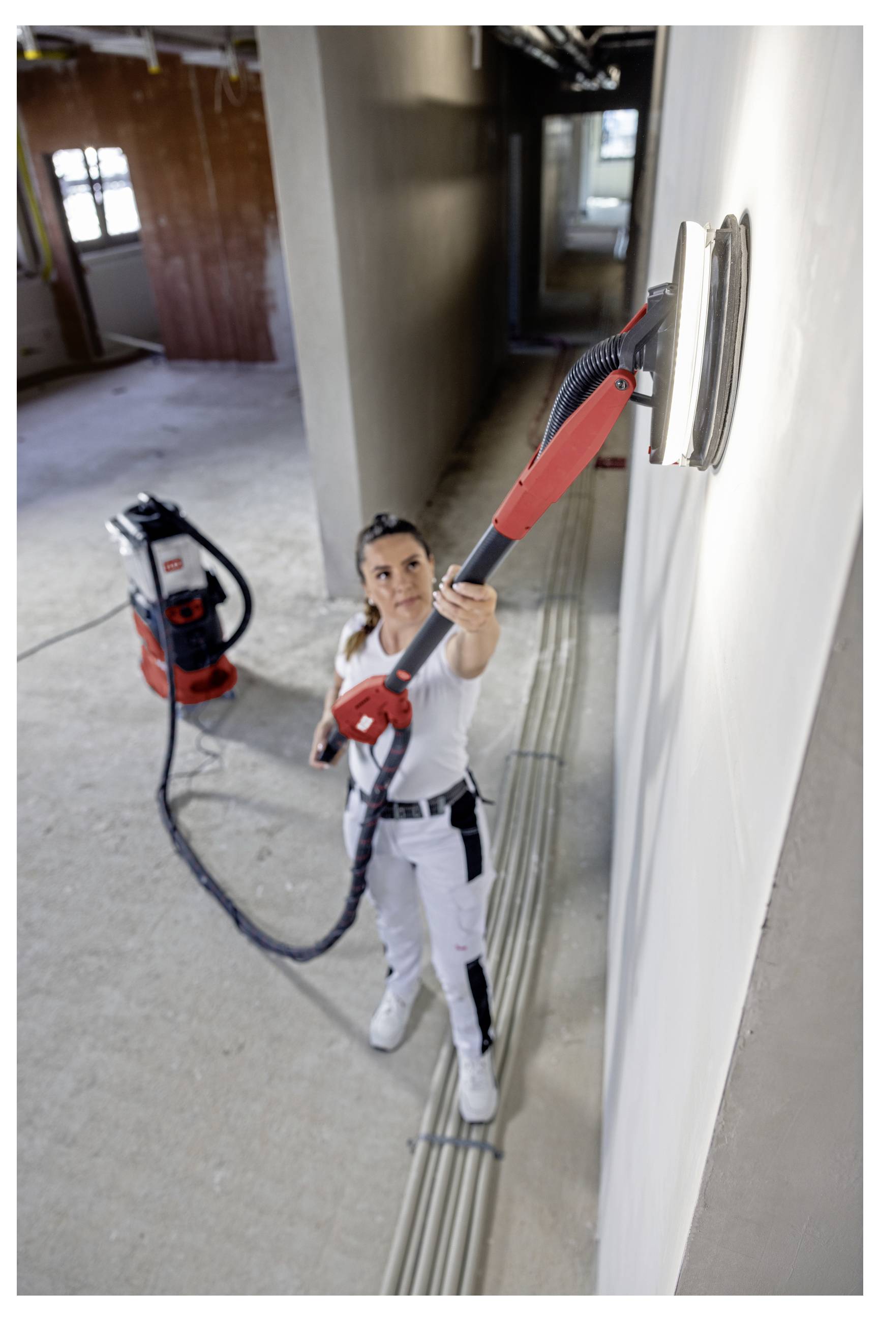 Arbeiter in weißer Schutzausrüstung, der eine Wand mit einem Elektroschleiffer mit Stange abschleifen, der an einen Absaugschlauch angeschlossen ist, in einem unfertigen Raum.