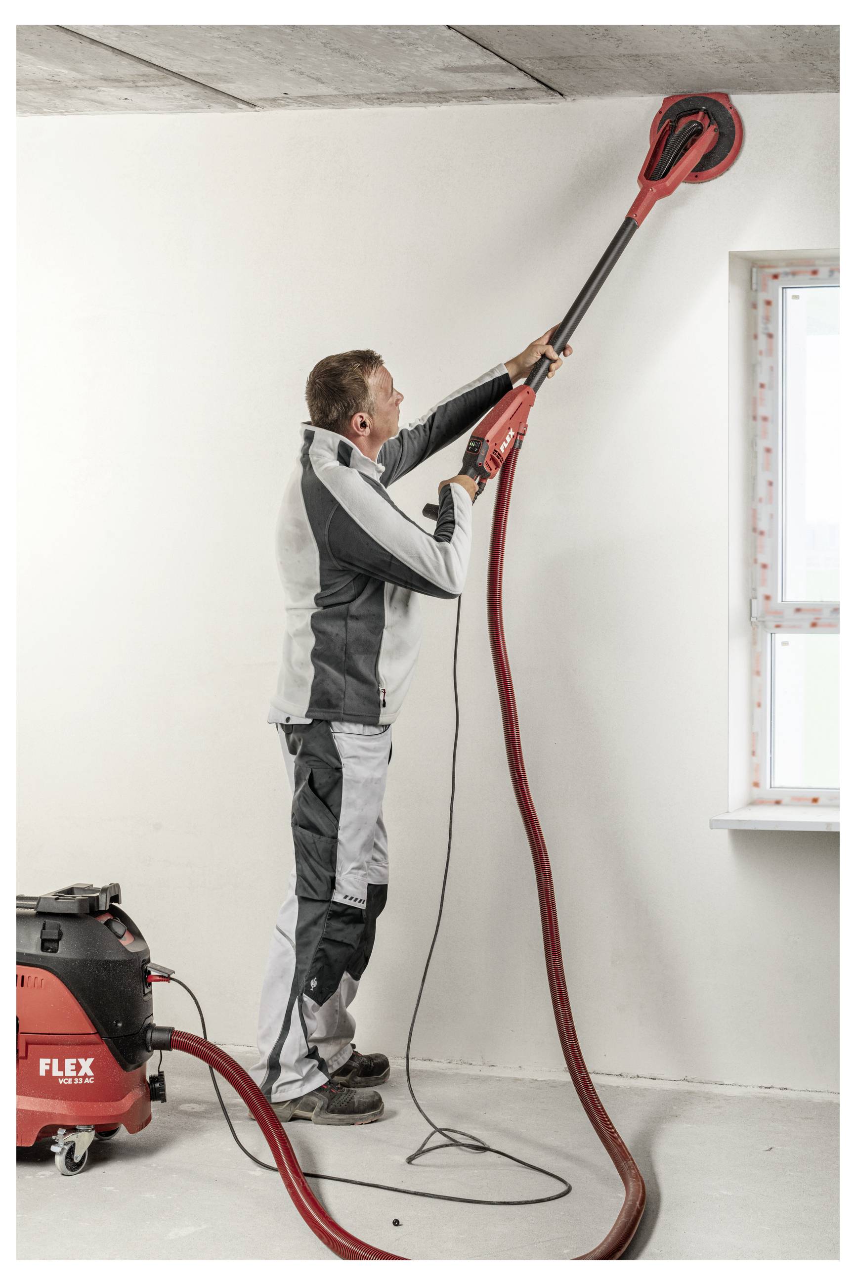 Arbeiter, der mit einem elektrischen Langschliff-Schleifers an einer Decke in einem renovierten Zimmer schleifen, mit einem Staubabzugsgerät neben ihm.