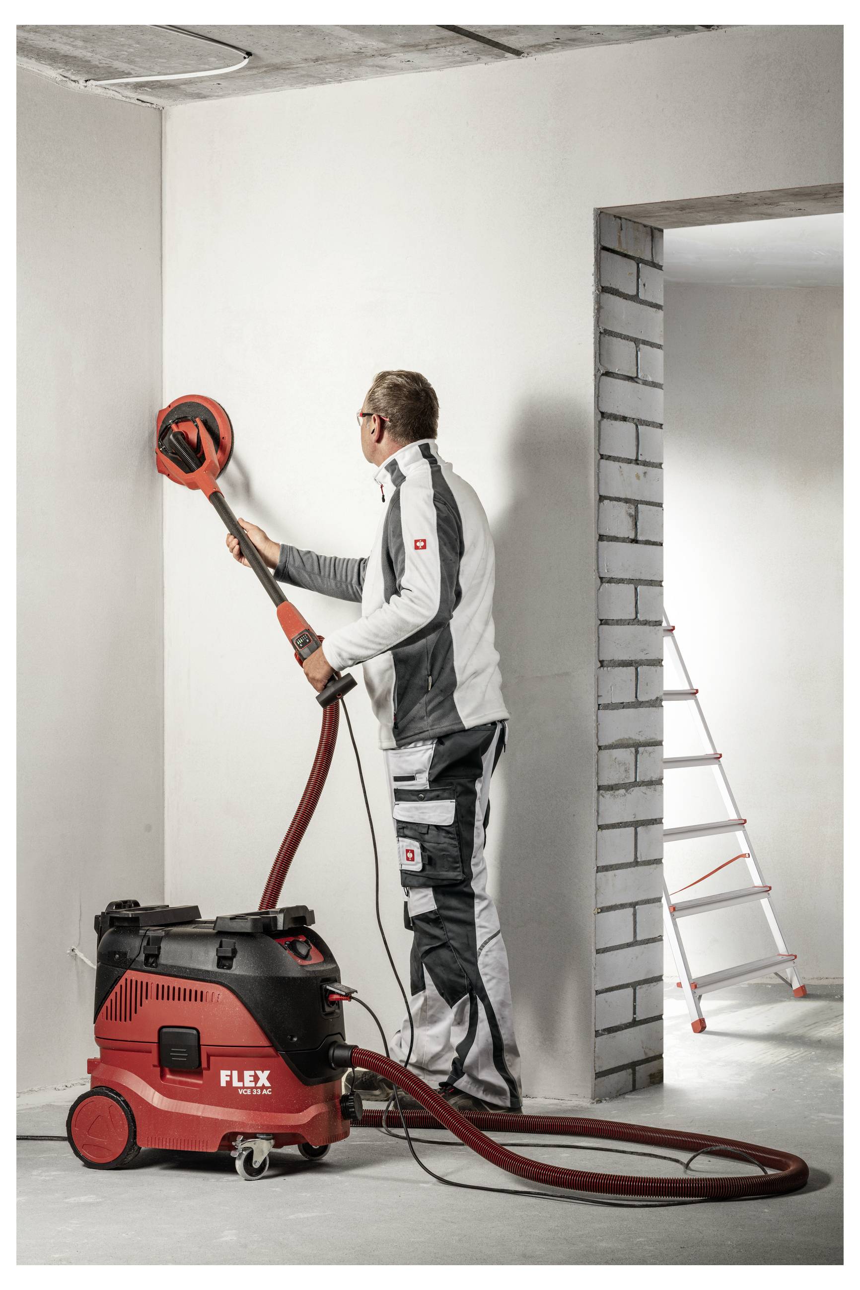 Arbeiter mit einem roten Wandschleifer, der an einem Staubsauger angebracht ist, in einem unvollendeten Raum, mit einer Leiter, die durch die Tür sichtbar ist.
