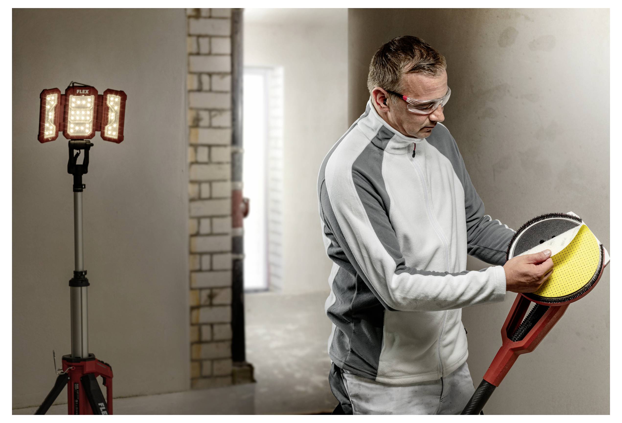 Ein Arbeiter in einer Schutzbrille inspiziert ein rotes Werkzeug oder eine rote Vorrichtung in Innenräumen und steht neben einer Stativ-Arbeitsleuchte in einem teilweise fertiggestellten Raum.