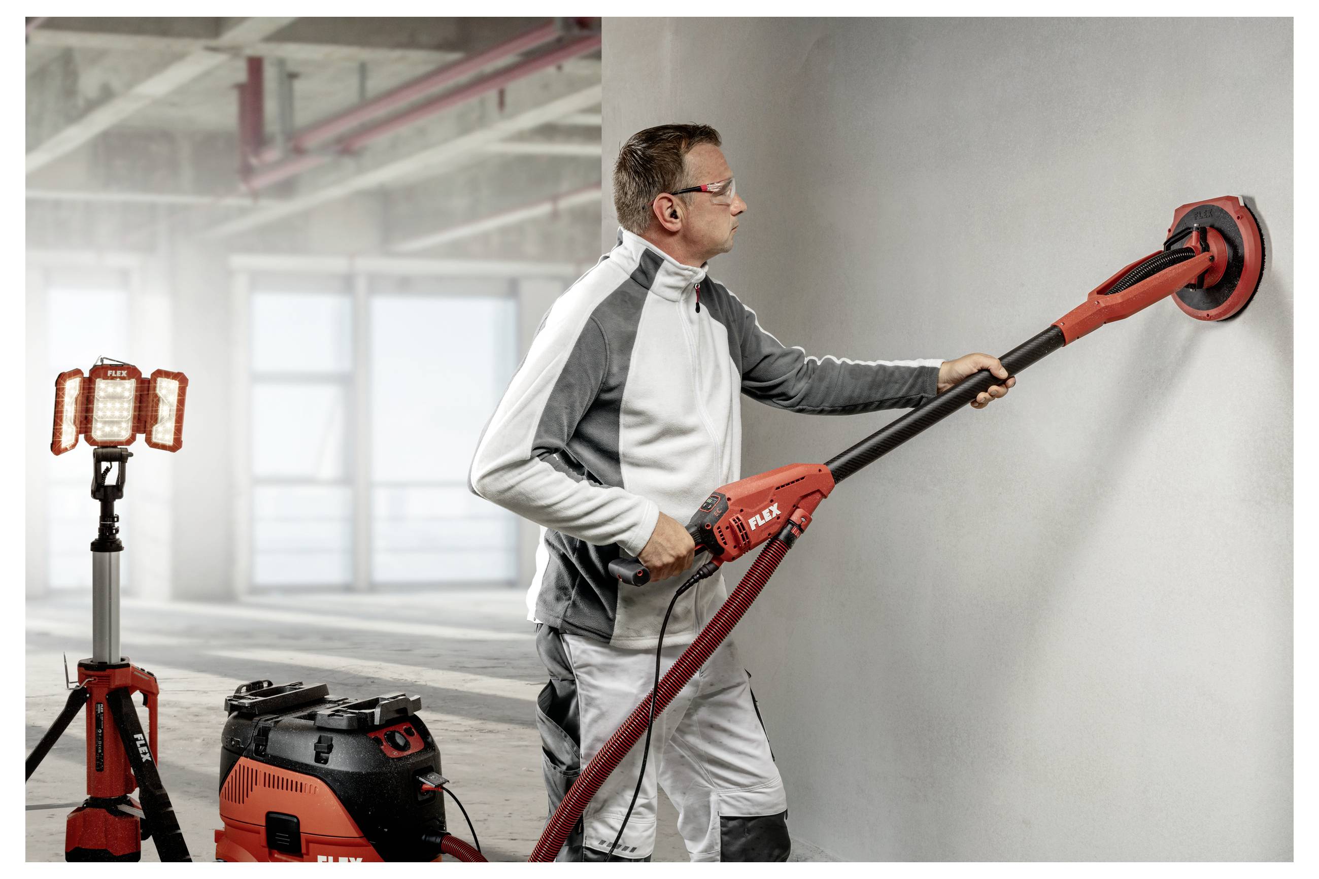 Ein Arbeiter verwendet einen rot-schwarzen Wandschlifer mit langem Griff an einer grauen Wand in einem Bauraum, mit einer tragbaren Arbeitsleuchte und einem Staubsauger in der Nähe.