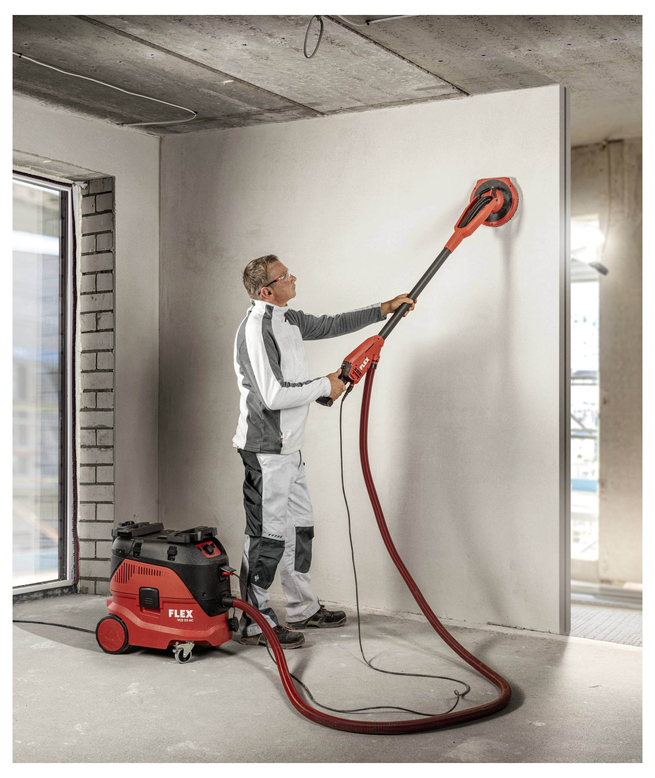 Arbeiter verwendet einen roten Trockenbau-Elektroschlifer mit langem Griff, um eine Innenwand zu glätten, mit einer Absauganlage auf dem Boden und in einer offenen Baustelle.