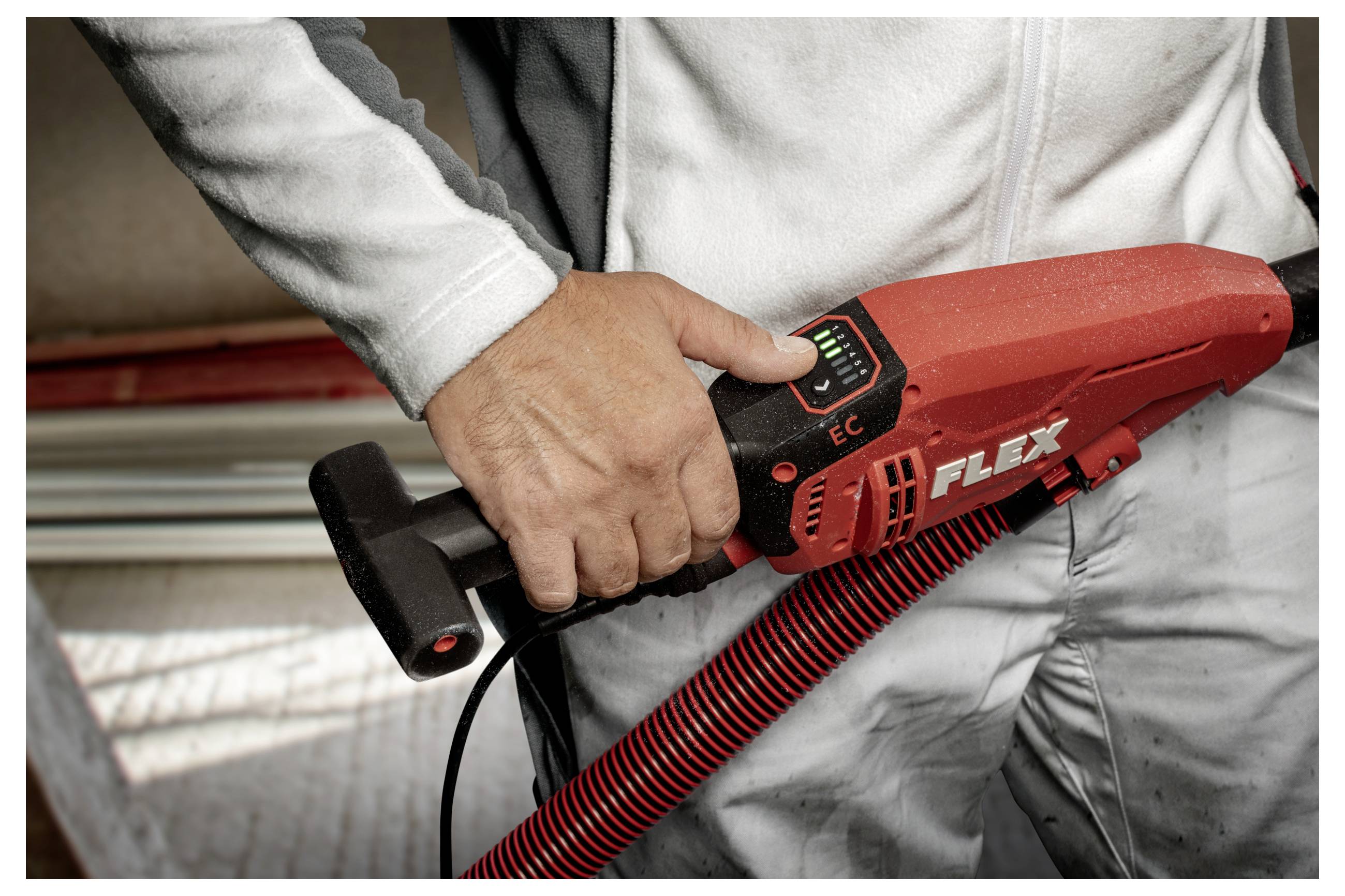 Arbeiter hält ein rotes FLEX-Elektrowerkzeug mit Griff und Stromkabel, in Nahaufnahme vor Arbeitsbekleidung dargestellt.