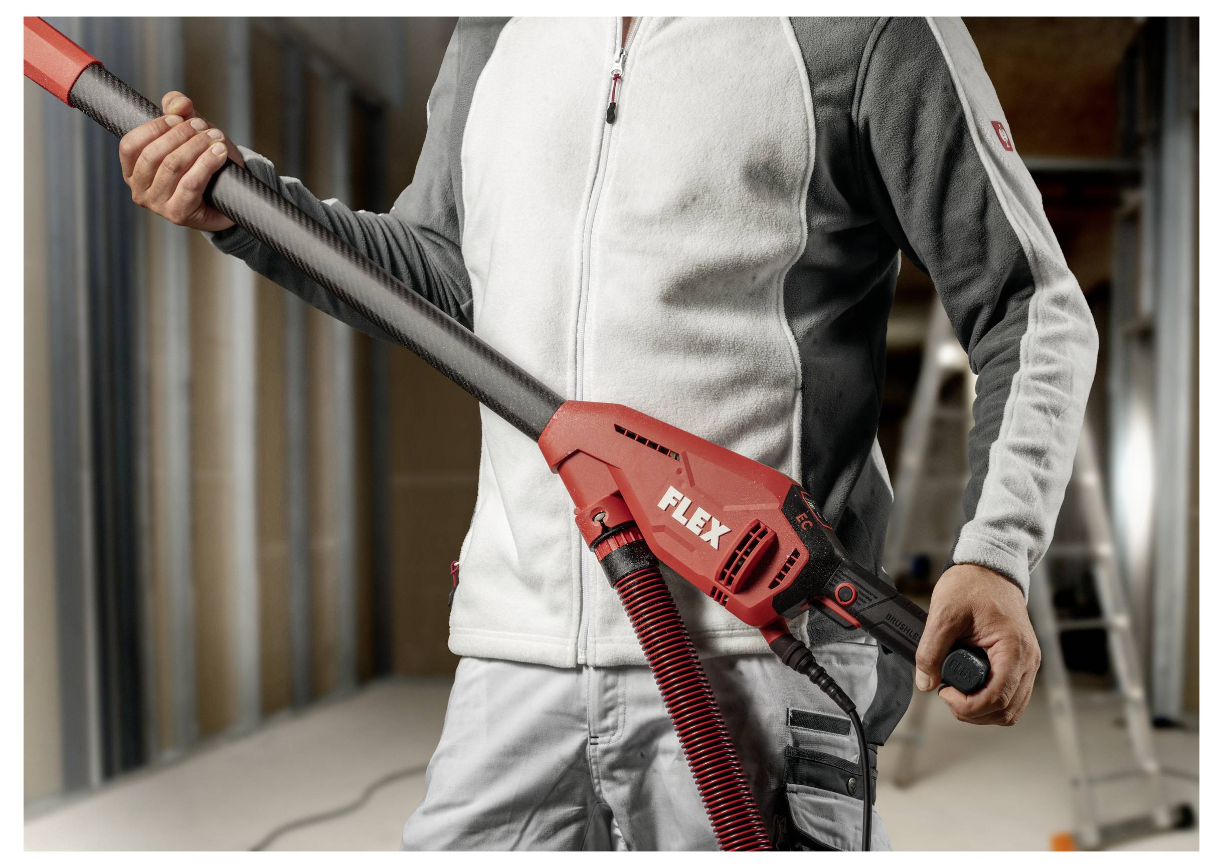 Arbeiter, der einen roten FLEX-Trockenbauschleifer mit langem Griff und Absaugschlauch auf einer Baustelle hält.