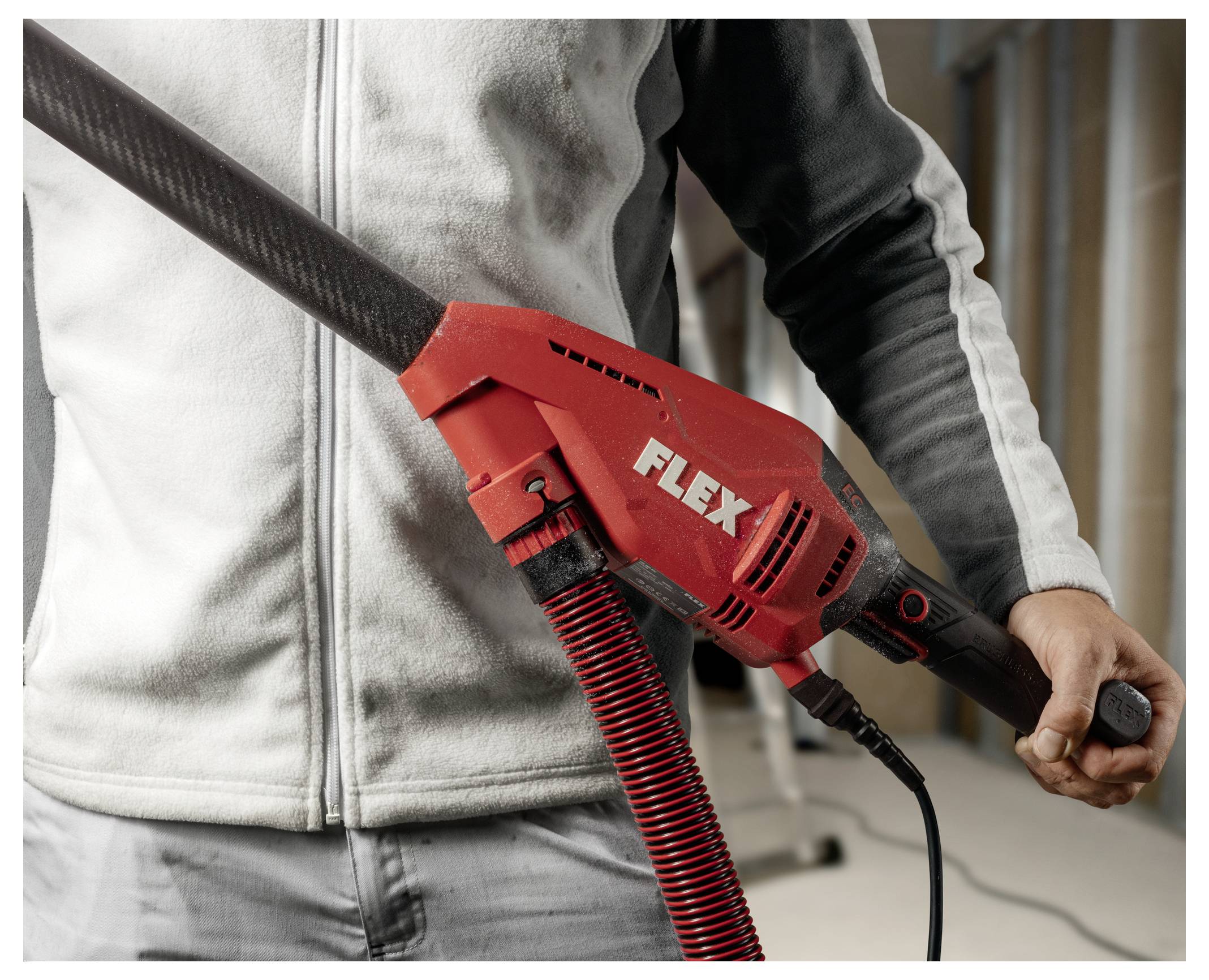 Person mit einem roten FLEX-Elektrowerkzeug mit einem langen angeschlossenen Schlauch oder Absaugschlauch, quer über dem Oberkörper gehalten, in einem Innenarbeitsbereich.
