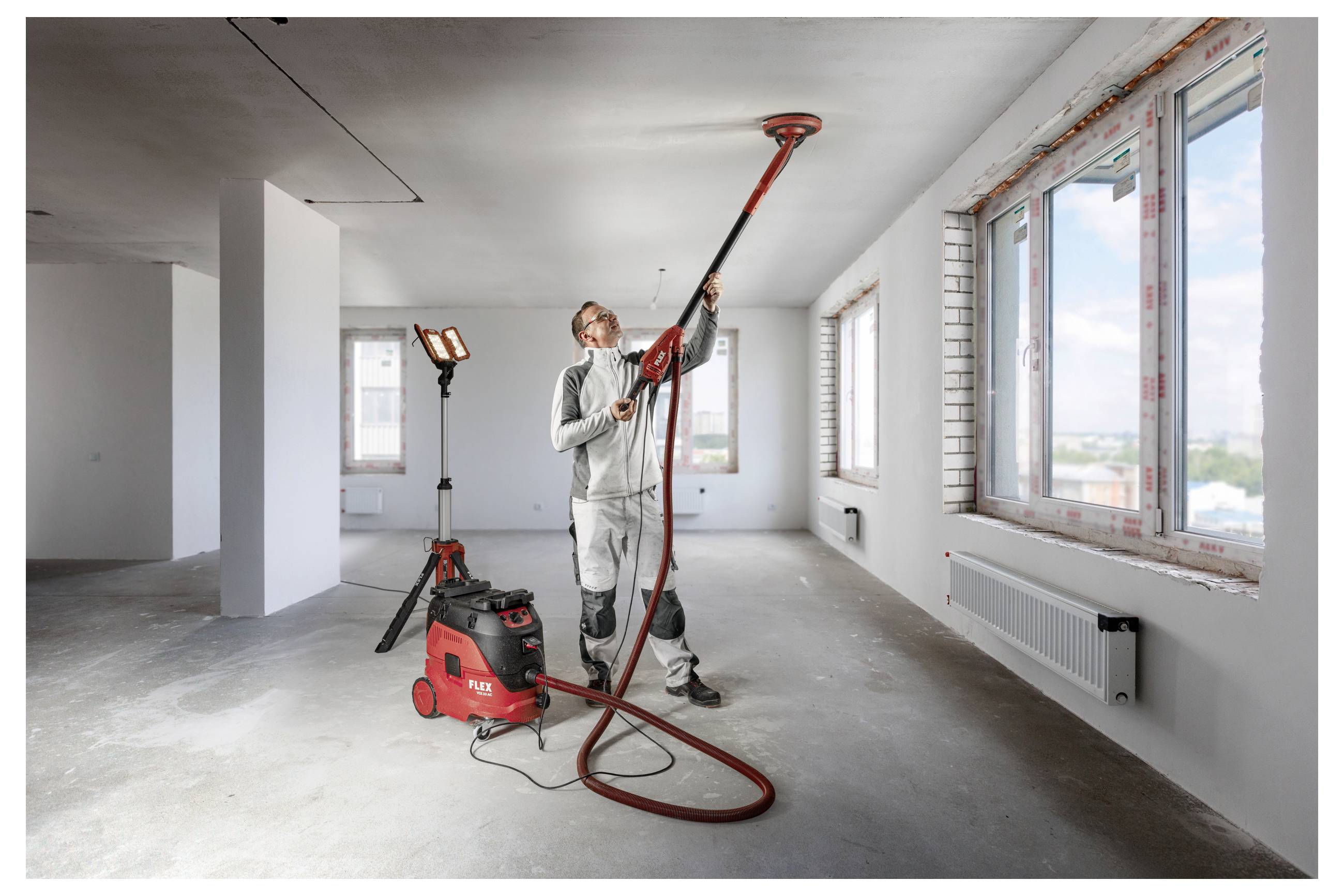 Arbeiter, der eine Decke mit einem Teleskopschleiffer in einem unfertigen Raum abschleidet, mit einem Staubabscheider und Fenstern entlang der Wand.