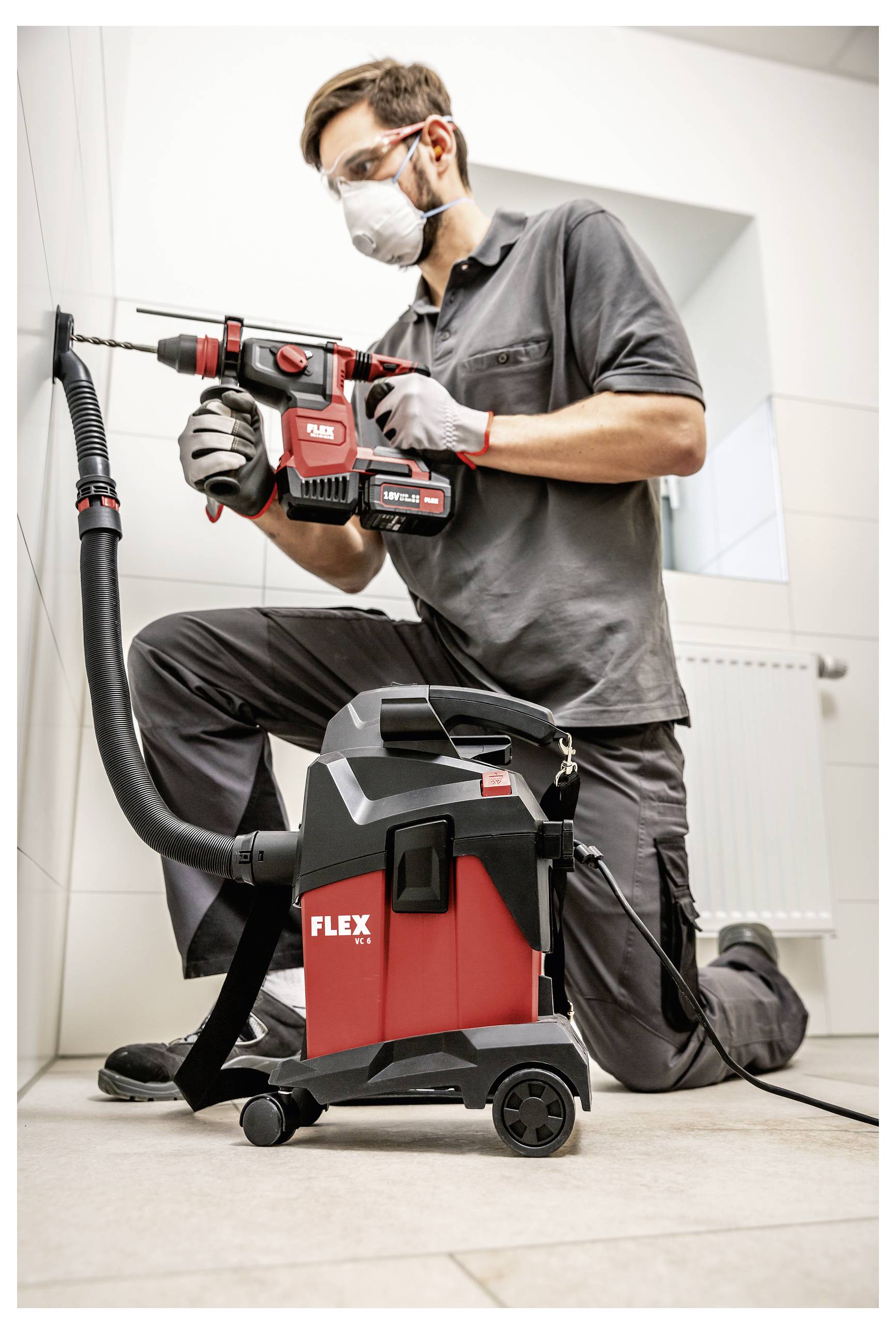 Arbeiter mit Maske, der eine rote FLEX-Bohrmaschine mit angeschlossener Absauganlage verwendet, während er drinnen kniet.