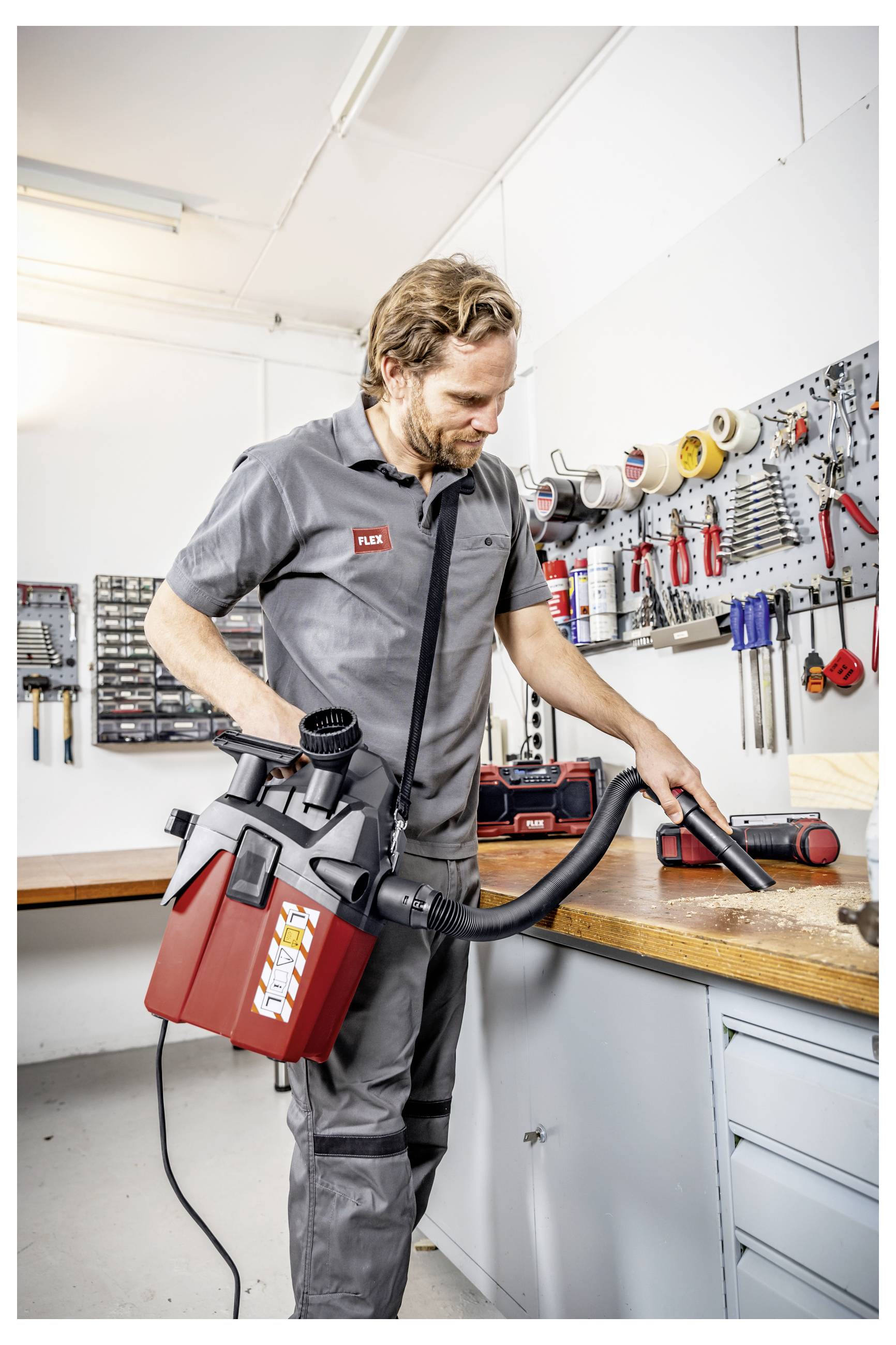 Ein Mechaniker benutzt einen tragbaren Nass-Trocken-Staubsauger zum Reinigen einer Werkbank in einer Werkstatt, mit Werkzeugen und Materialien, die an der Wand hinter ihm organisiert sind.
