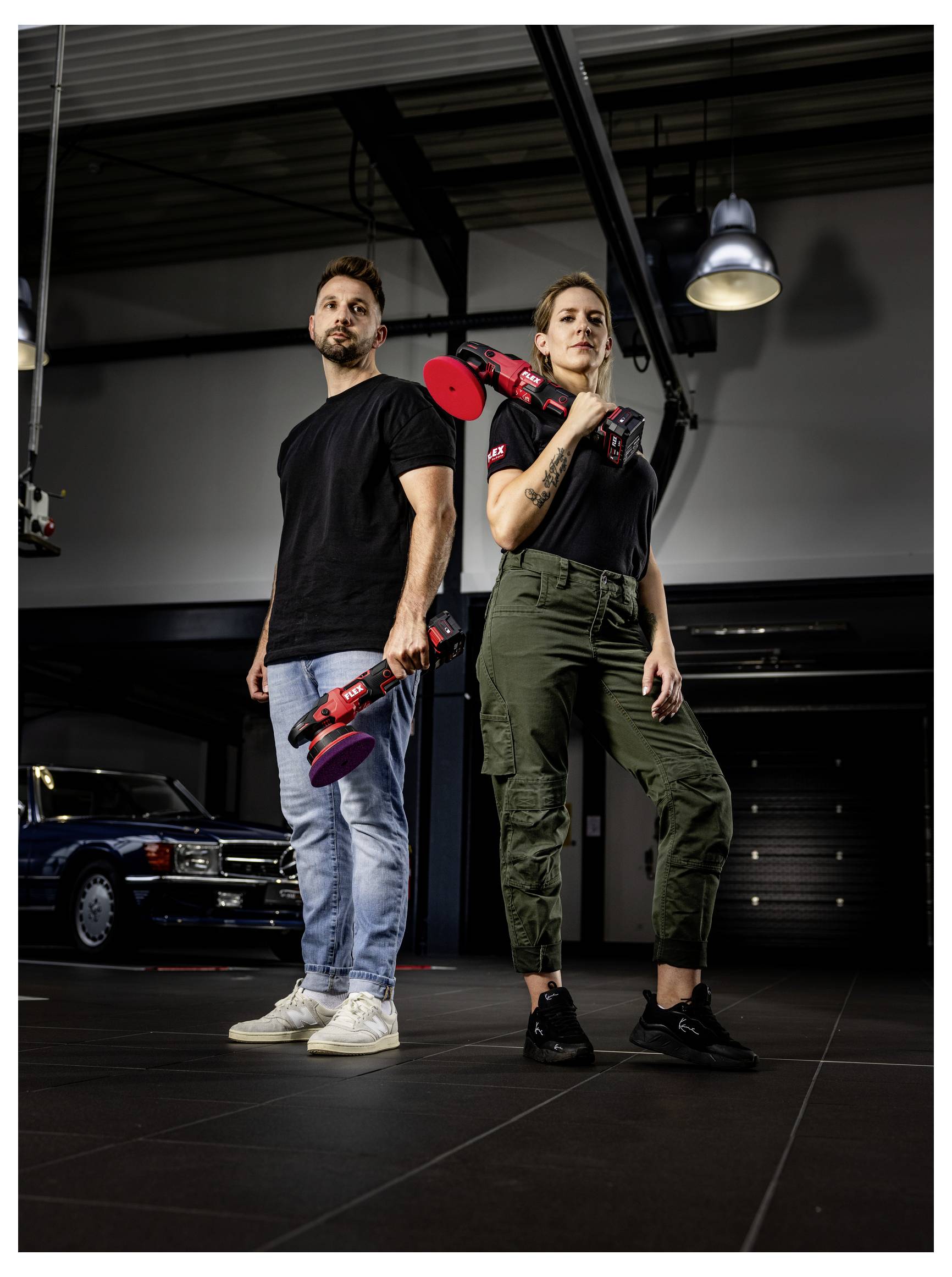 Zwei Personen stehen in einer Garage und halten rote Elektrowerkzeuge, während sie Rücken an Rücken unter industrieller Beleuchtung posieren.