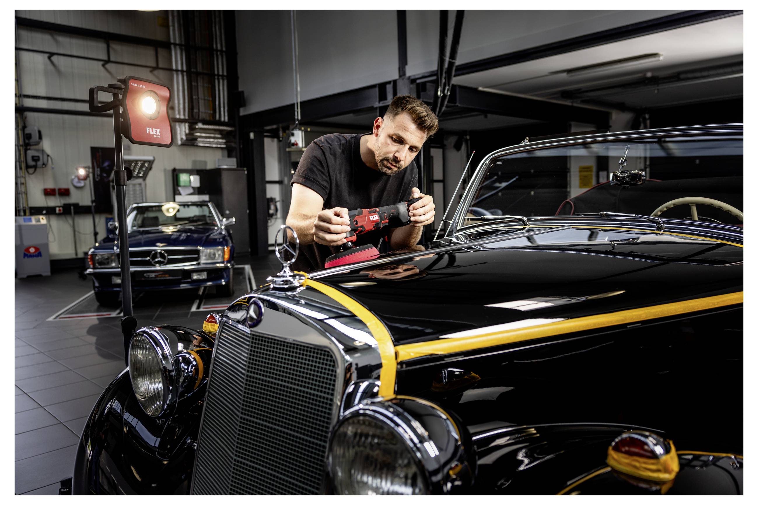 Ein Mechaniker poliert die Motorhaube eines schwarzen Vintage-Mercedes in einer Werkstatt, mit einem weiteren Klassiker und Werkstattausrüstung im Hintergrund.