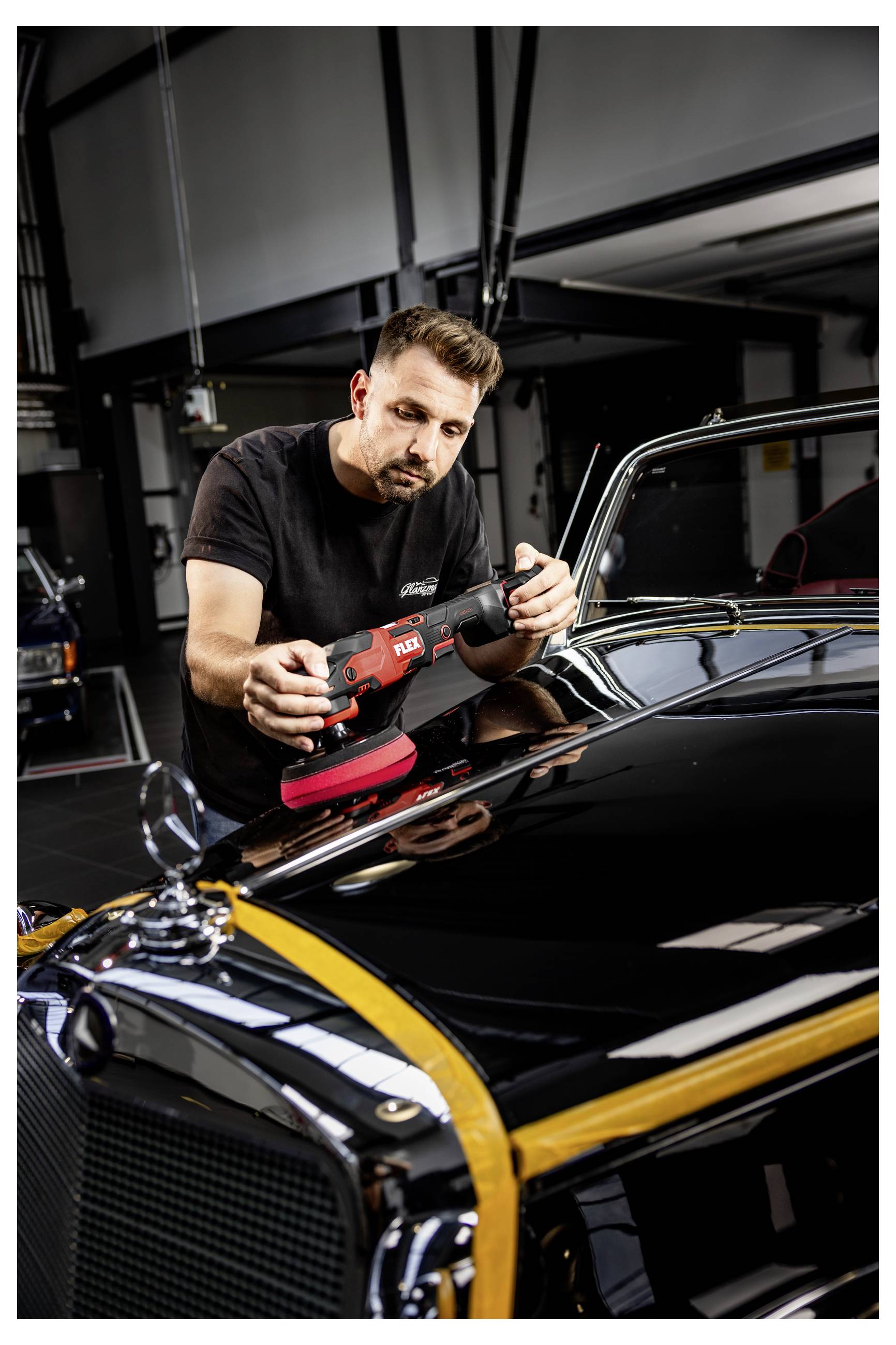 Ein Mann poliert ein schwarzes Klassikauto mit einem tragbaren Poliergerät in einer Garage und konzentriert sich auf die Motorhaube und die Chromverkleidung.