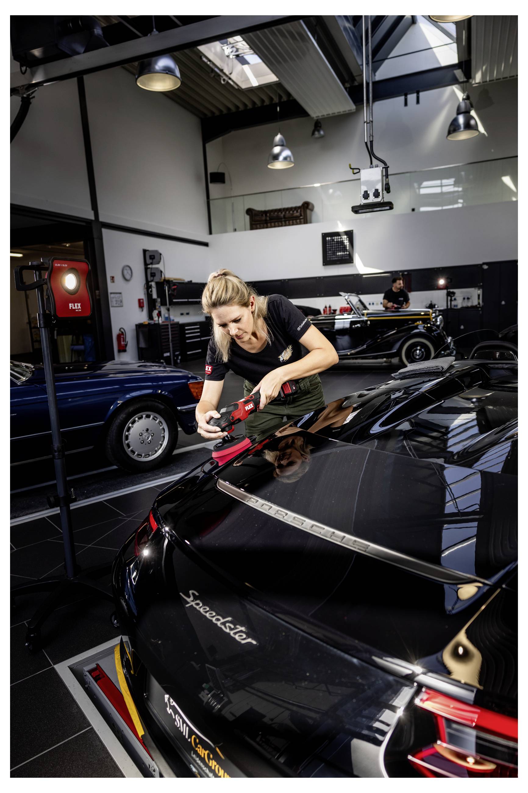 Frau poliert einen schwarzen Porsche Speedster in einem Autoausstellungsraum, mit anderen Klassikern im Hintergrund.