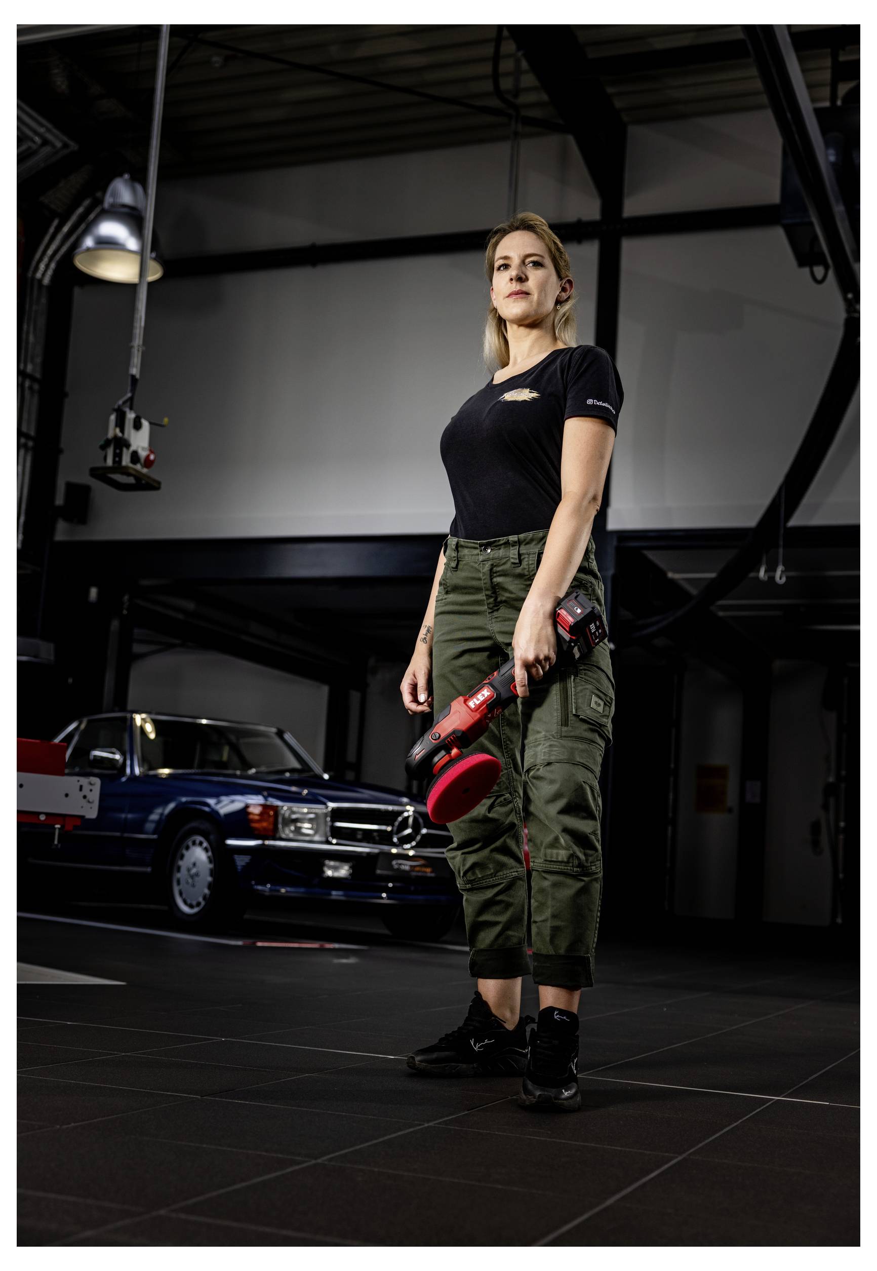Eine Frau steht in einer Garage oder Werkstatt und hält ein rotes Elektrowerkzeug, während im Hintergrund ein klassisches Auto zu sehen ist.