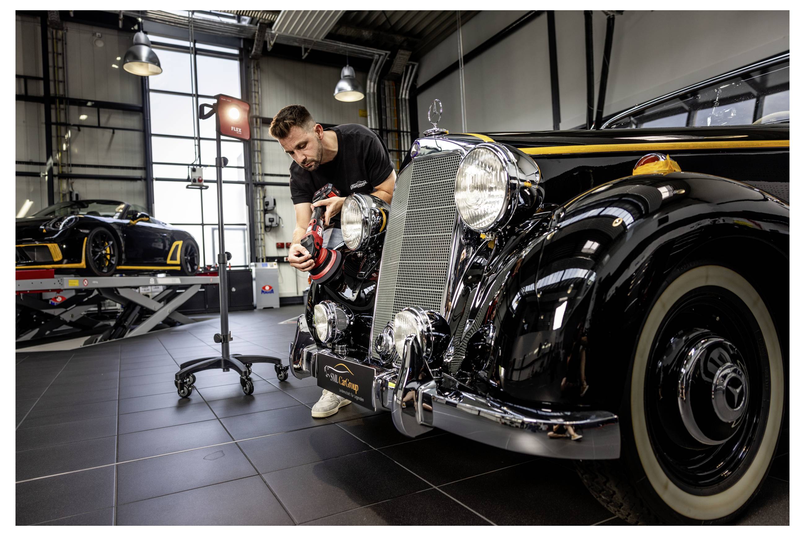Ein Mechaniker poliert das Scheinwerfer eines schwarzen Oldtimers in einer Werkstatt, während ein anderes Klassiker-Auto auf einer Hebebühne im Hintergrund steht.