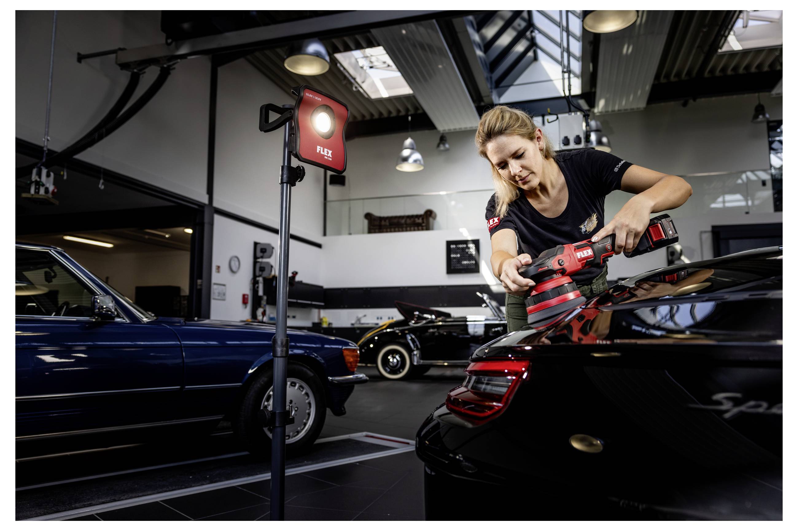 Frau poliert das Heck eines schwarzen Porsche-Sportwagens in einer Auto-Detailing-Werkstatt, mit anderen klassischen Autos im Hintergrund.