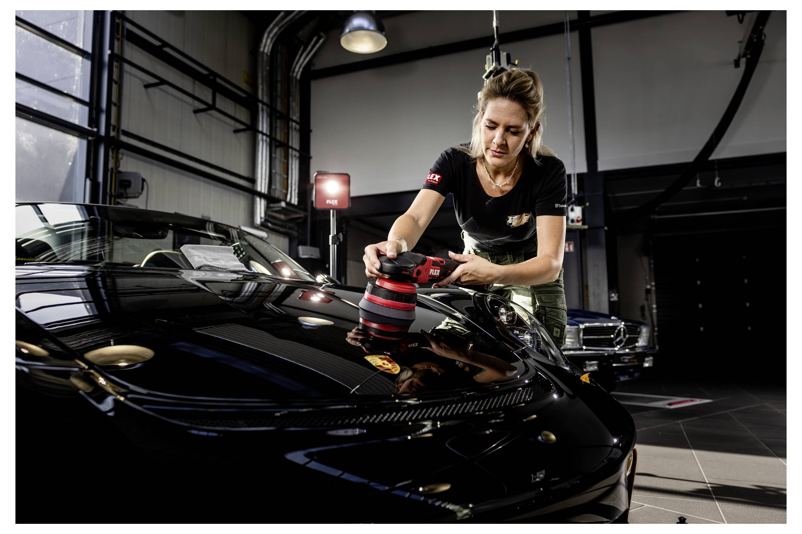 Frau, die mit einem tragbaren Poliergerät die Motorhaube eines schwarzen Autos in einer Autowäsche-Werkstatt poliert.