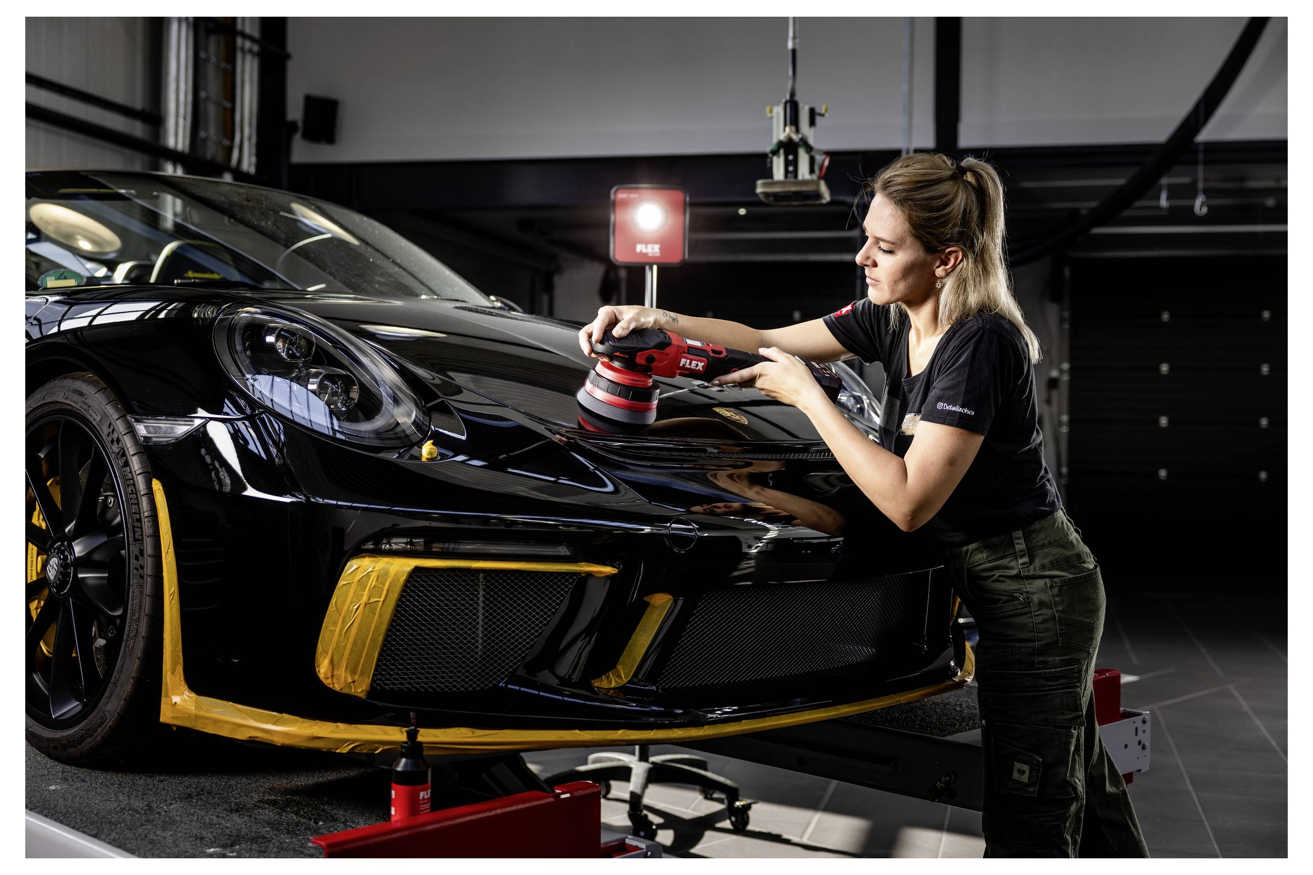 Frau poliert die Motorhaube eines schwarzen Sportwagens in einer Werkstatt mit einem roten elektrischen Poliergerät.