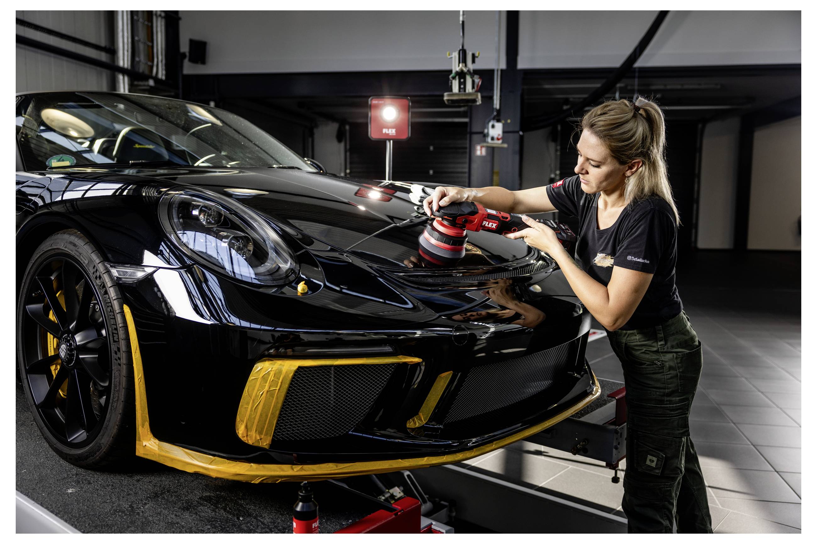 Frau poliert die Motorhaube eines schwarzen Sportwagens in einer Garage und benutzt dabei einen roten Poliermaschine, während das Auto auf einer Hebebühne angehoben ist.