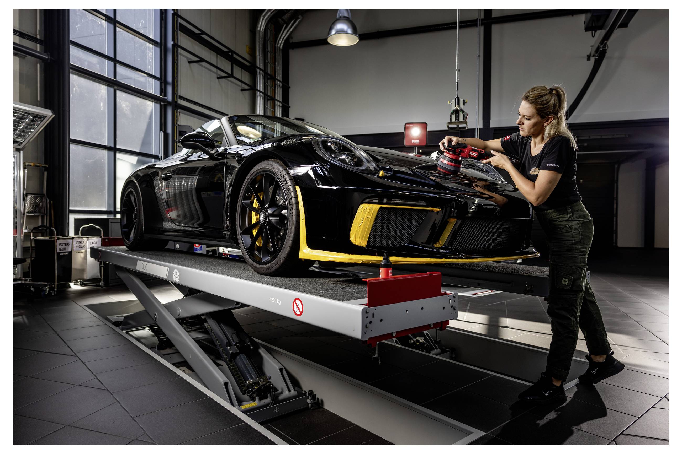 Techniker poliert die Vorderseite eines schwarzen Sportwagens auf einer Hydraulikbühne in einer modernen Garage oder in einem Detailing-Shop.