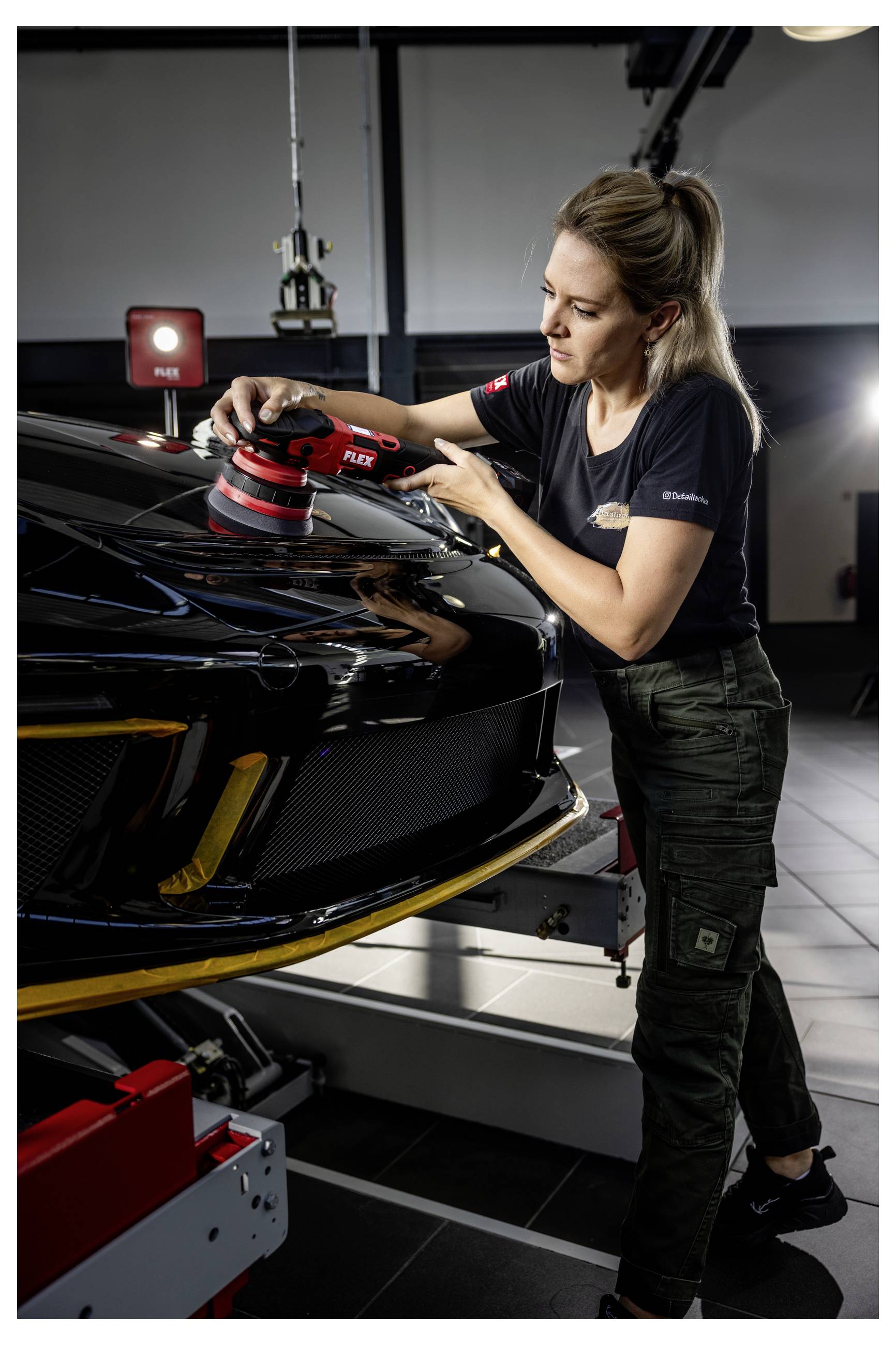 Frau poliert die Motorhaube eines schwarzen Sportwagens mit einem Elektroschleiffer in einer Garage oder Detailing-Werkstatt.