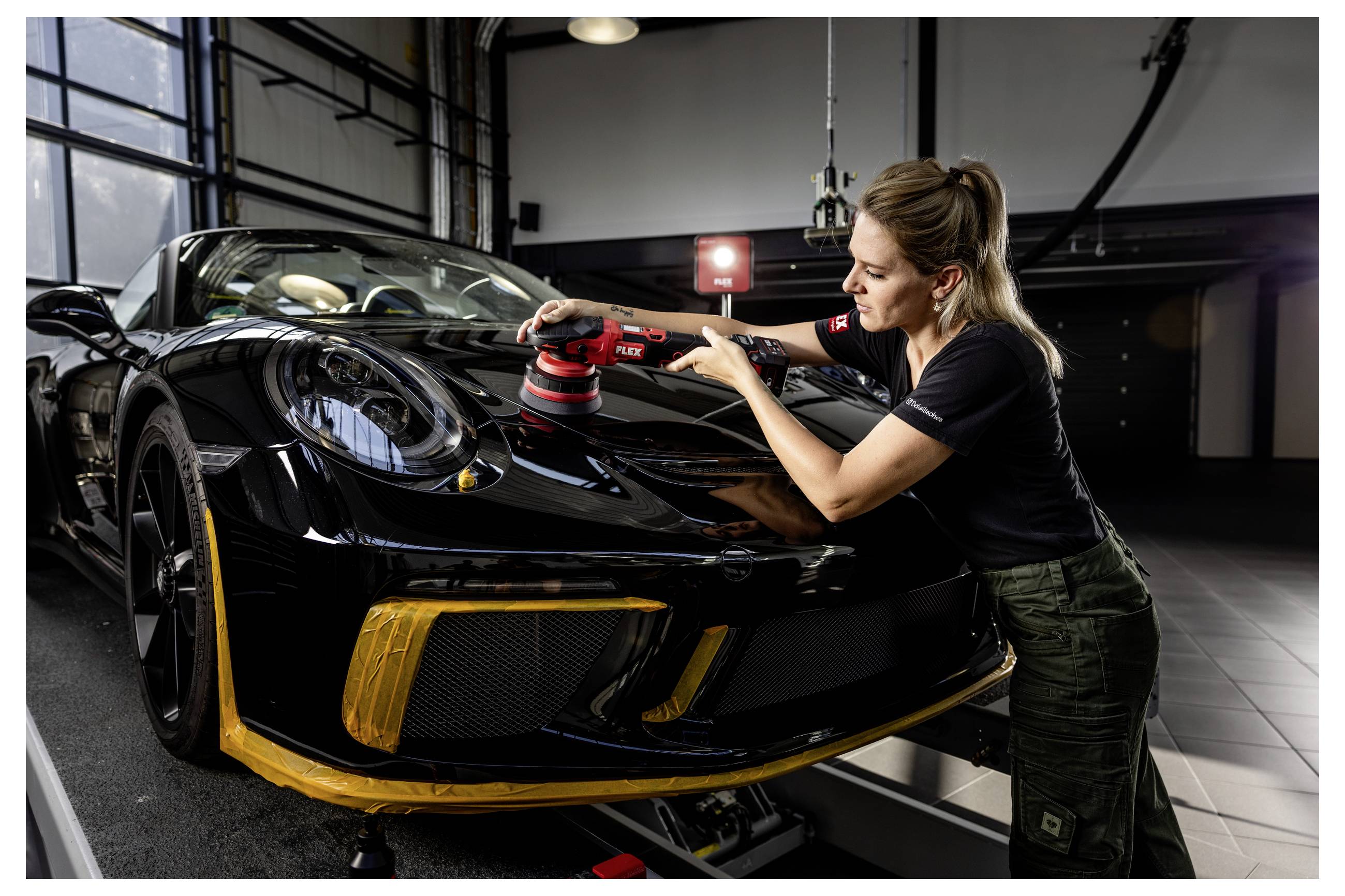 Frau poliert die Motorhaube eines schwarzen Sportwagens in einer Garage oder Detailingwerkstatt mit einem Poliergerät.