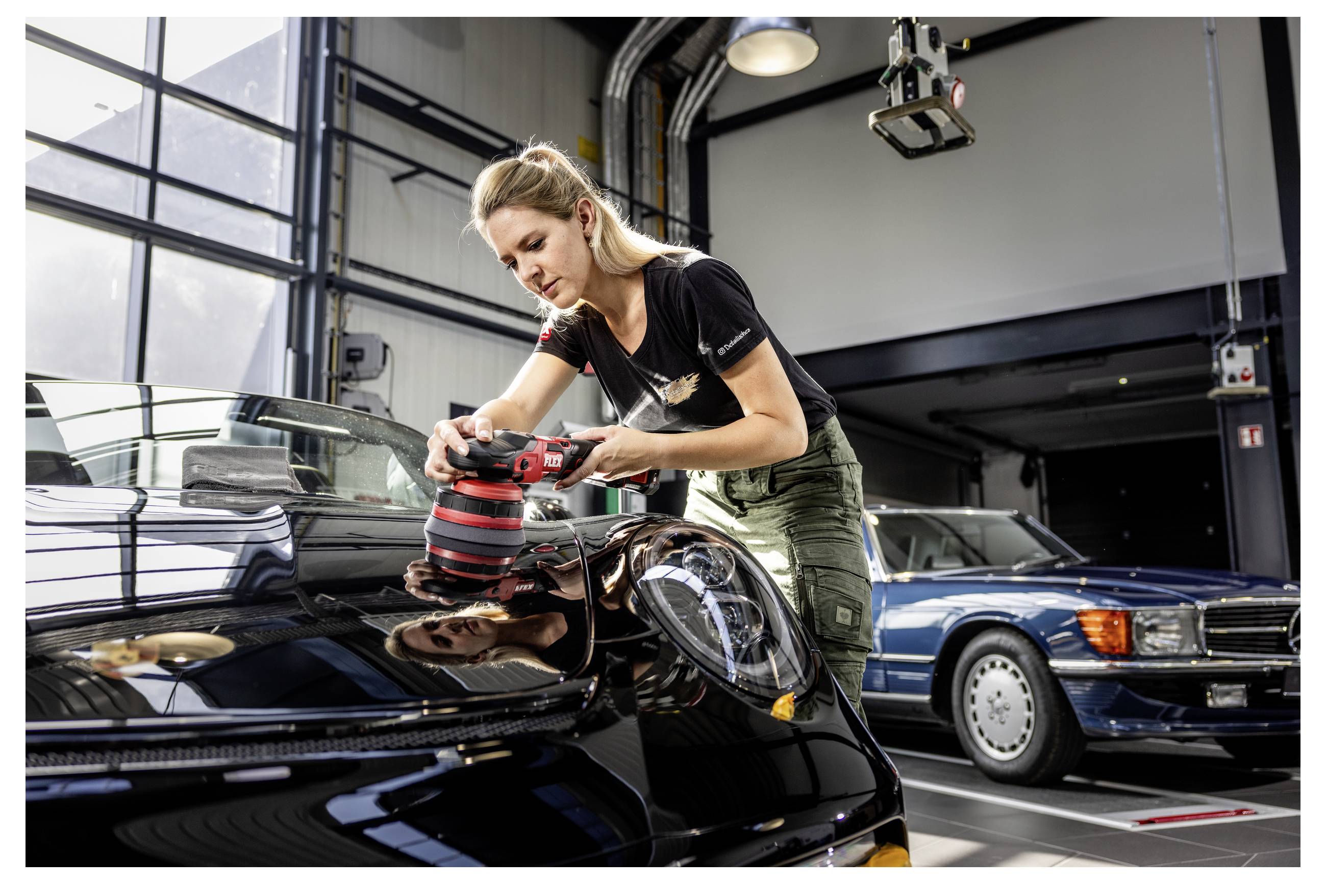 Frau poliert die Motorhaube eines schwarzen Autos in einer Garage, mit einem anderen klassischen Auto, das im Hintergrund geparkt ist.