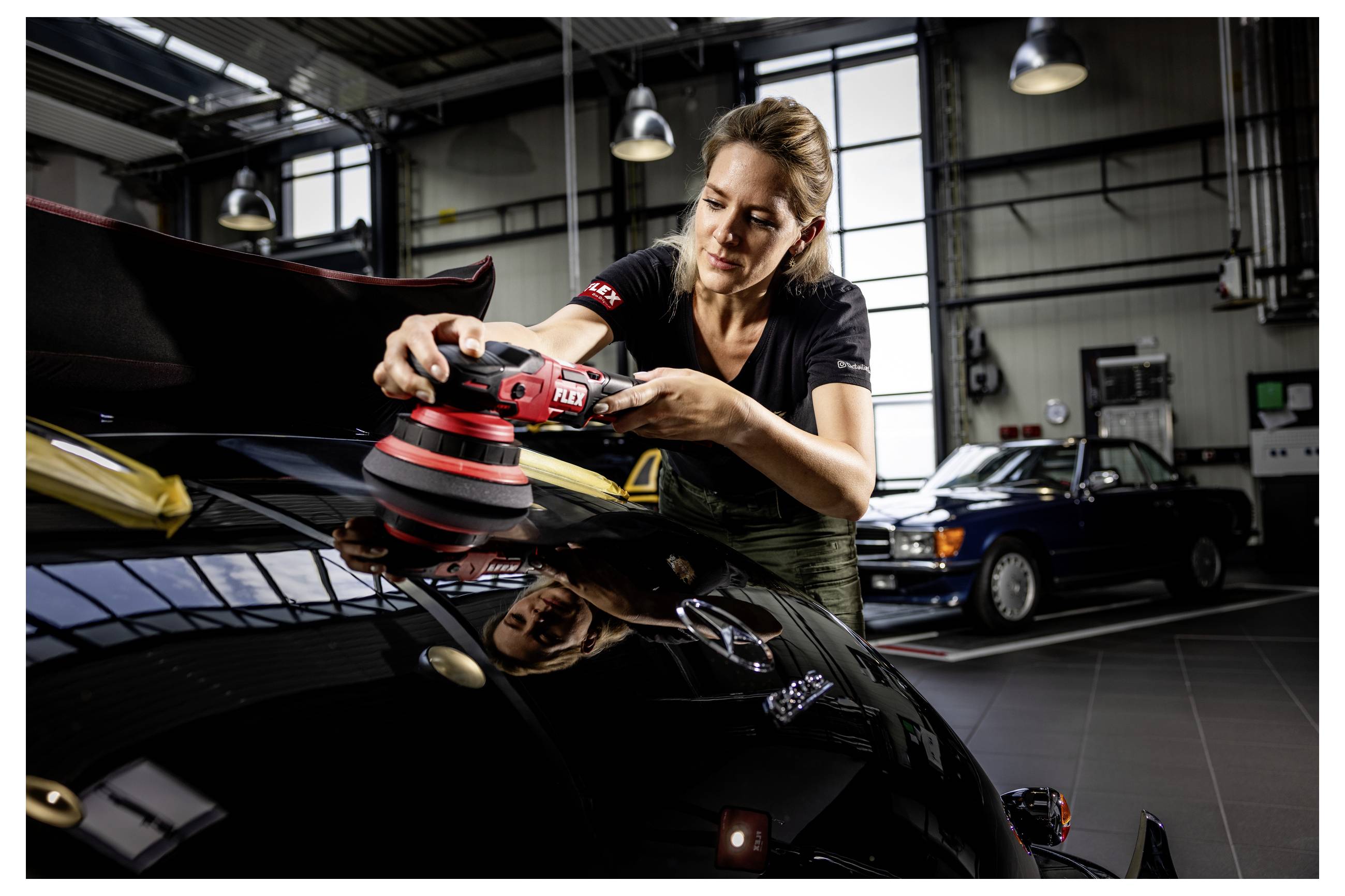 Eine Frau poliert die Motorhaube eines schwarzen Sportwagens mit einem Elektroschleifer in einer Garage, während ein anderes Auto im Hintergrund zu sehen ist.