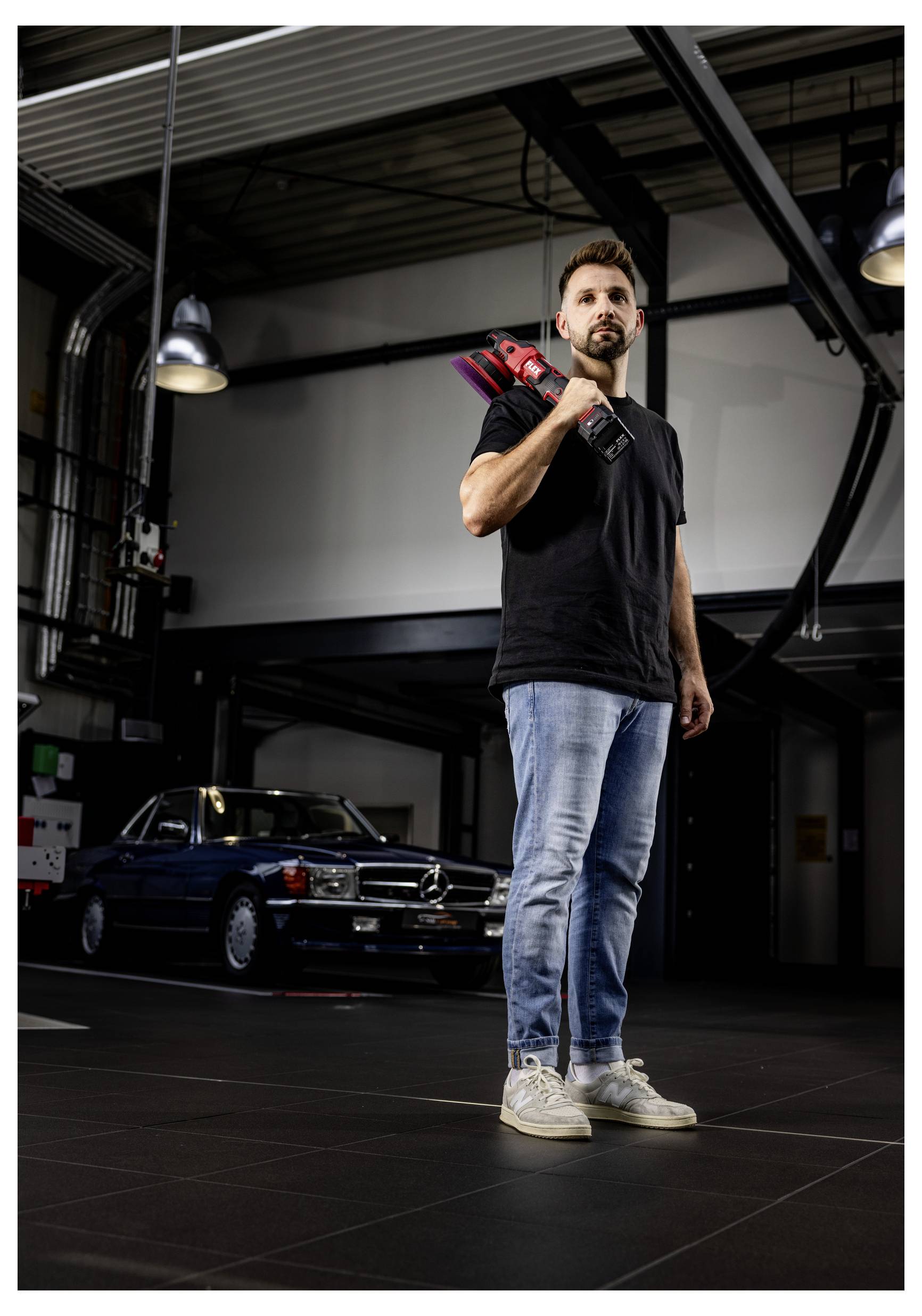 Ein Mann steht in einer Garage und hält ein Elektrowerkzeug auf seiner Schulter, während hinter ihm ein vintage schwarzes Auto geparkt ist.