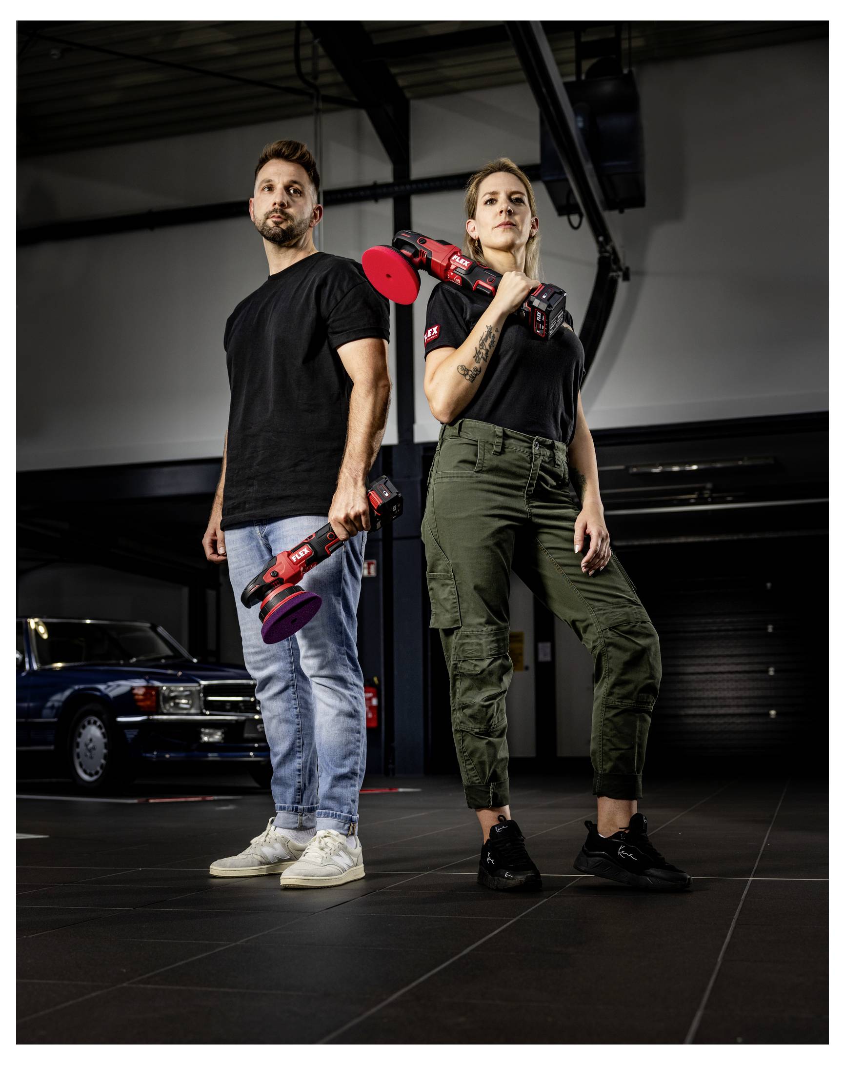 Zwei Mechaniker stehen in einer Werkstatt und halten rote Elektroschleifermaschinen, posiert Rücken an Rücken neben einem Klassiker.