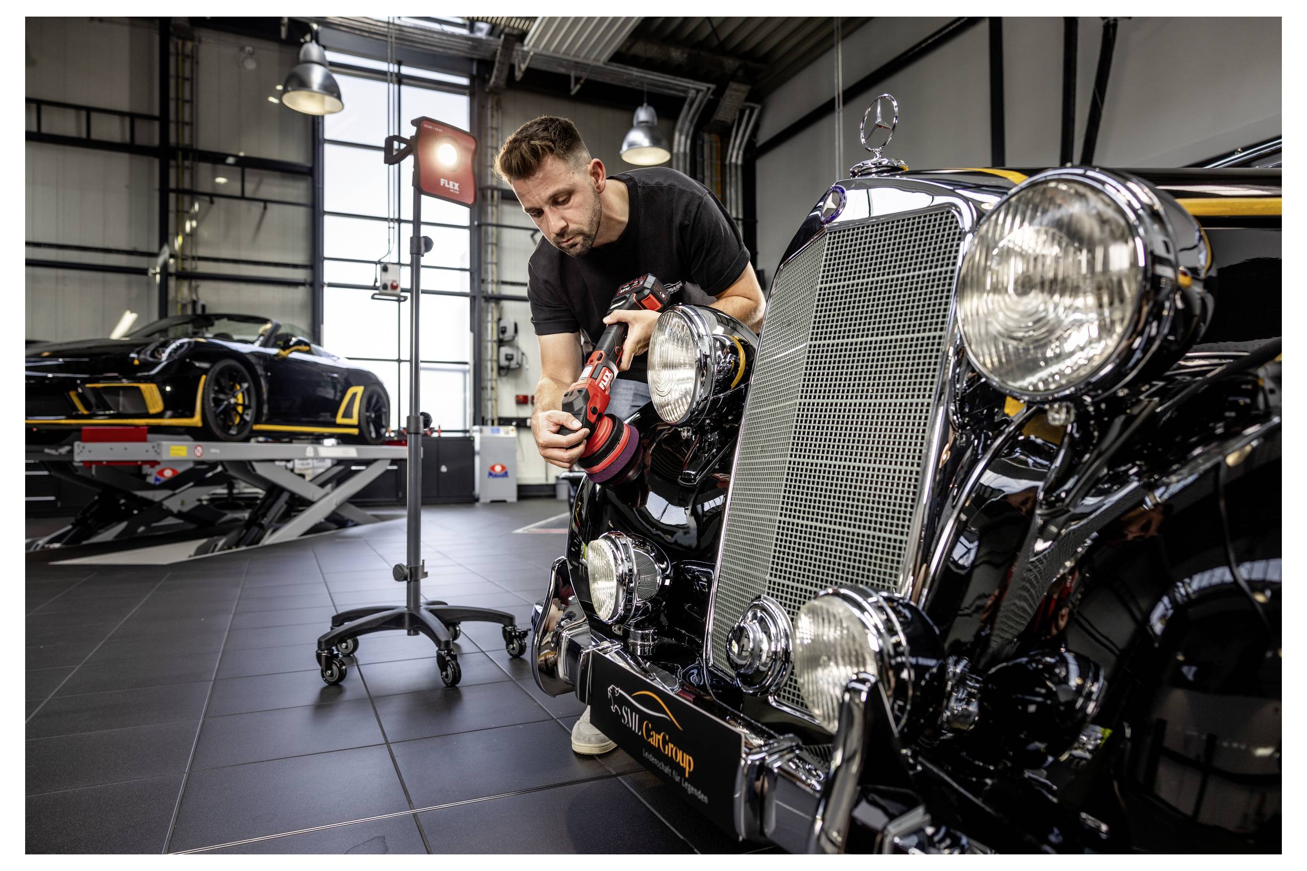 Ein Mechaniker poliert die Vorderseite eines klassischen schwarzen Autos in einer Werkstatt, während sich ein anderes Sportauto auf einer Hebebühne im Hintergrund befindet.
