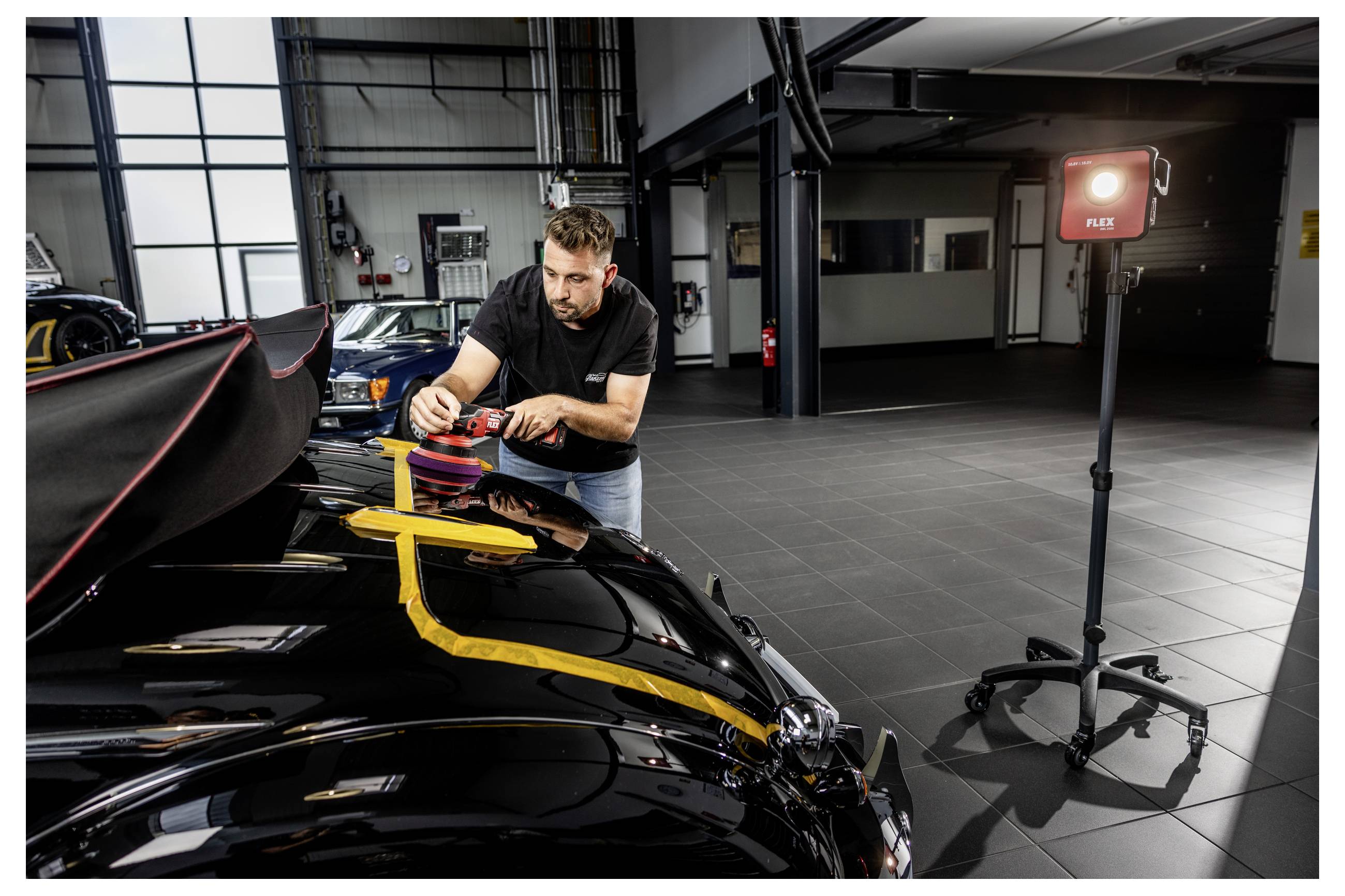 Ein Mann poliert mit einem Polierwerkzeug die Motorhaube eines schwarzen Autos in einer Garage, während in der Nähe ein rotes Arbeitslicht auf einem Ständer steht.