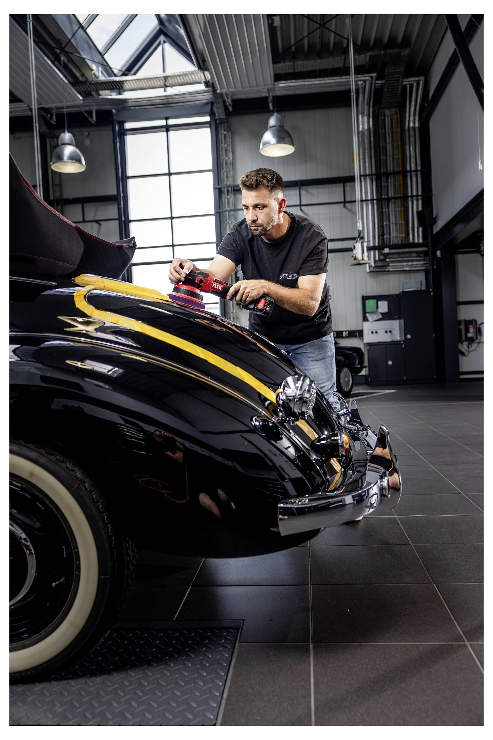 Ein Mann poliert ein schwarzes Vintage-Auto mit gelben Streifen in einer garageähnlichen Werkstatt.