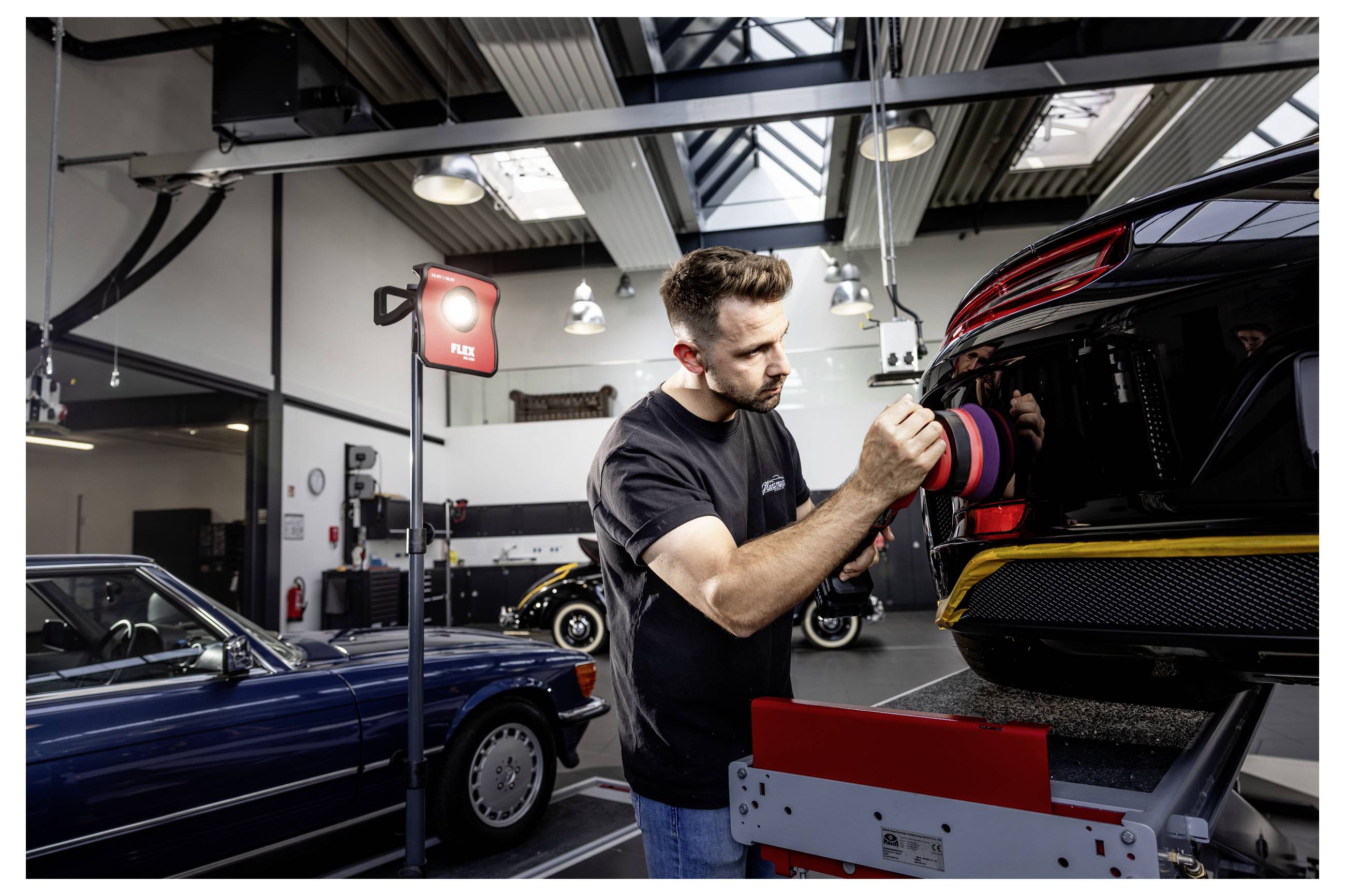 Ein Mechaniker poliert das Heck eines schwarzen Sportwagens mit einem Polierer in einer Autowäscherei, während andere Autos im Hintergrund zu sehen sind.