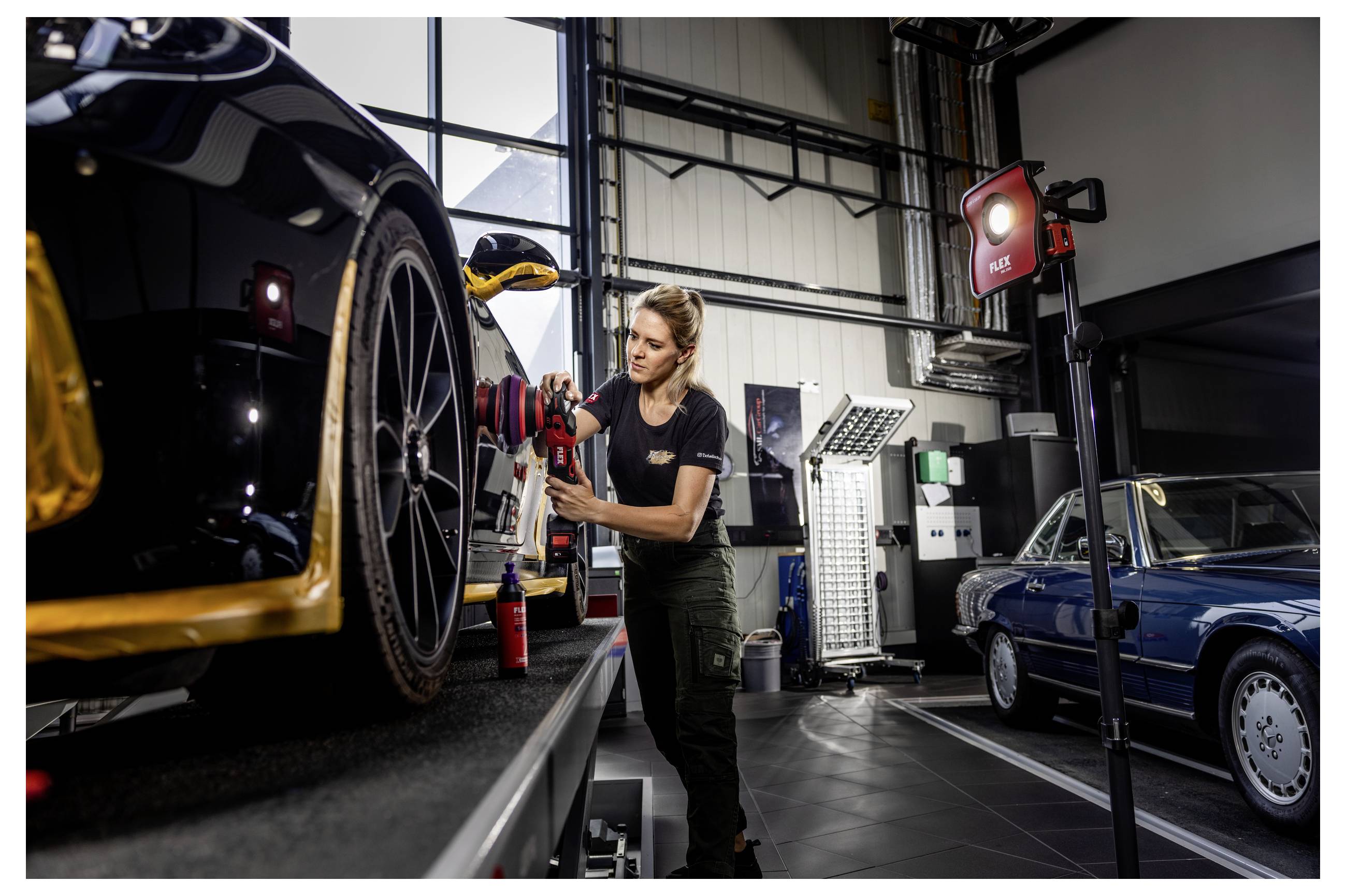 Ein Mechaniker stellt das Vorderrad und die Bremsanlage eines schwarzen Sportwagens in einer Garage ein, mit einem weiteren Klassiker und Werkstattleuchten im Hintergrund.