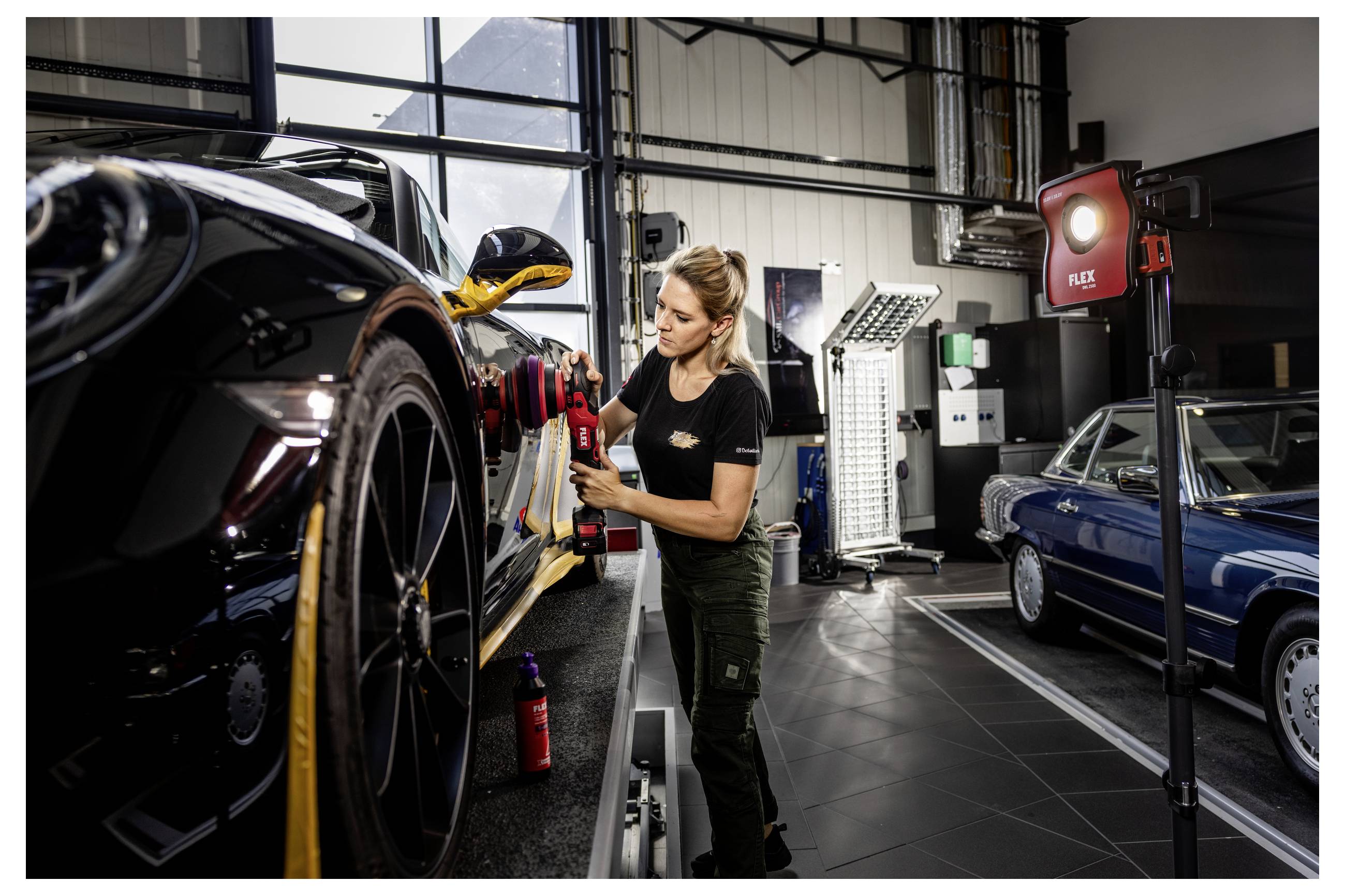 Ein Mechaniker poliert ein schwarzes Sportauto in einer Autowerkstatt, mit einem weiteren Klassiker und Arbeitsleuchten im Hintergrund.