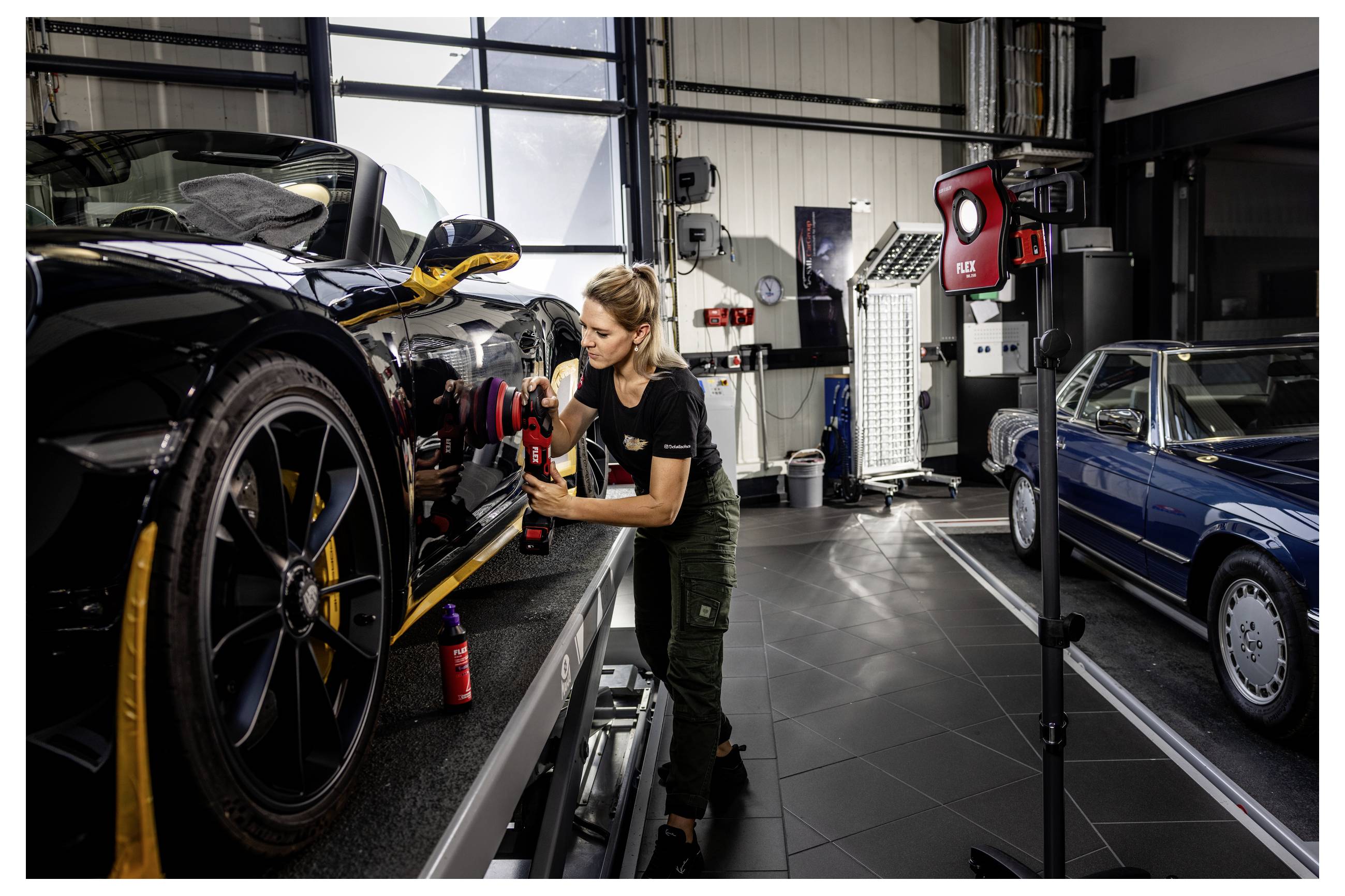 Ein Mechaniker poliert das Vorderrad eines schwarzen Sportwagens in einer Werkstatt, während ein anderes klassisches blaues Auto in der Nähe geparkt ist.