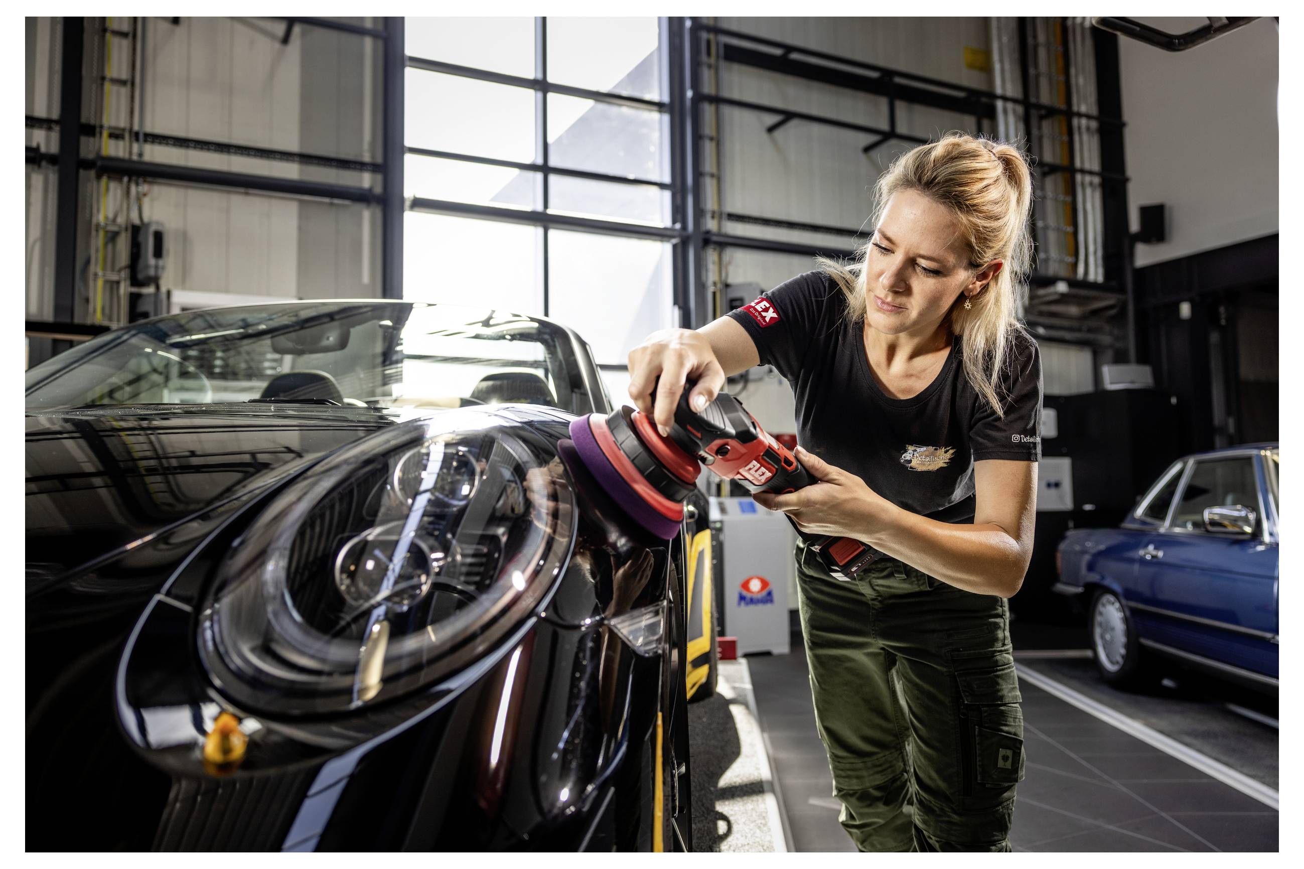 Eine Frau poliert die Vorderseite eines schwarzen Sportwagens mit einem Elektroschleiffer in einer Autowerkstatt.