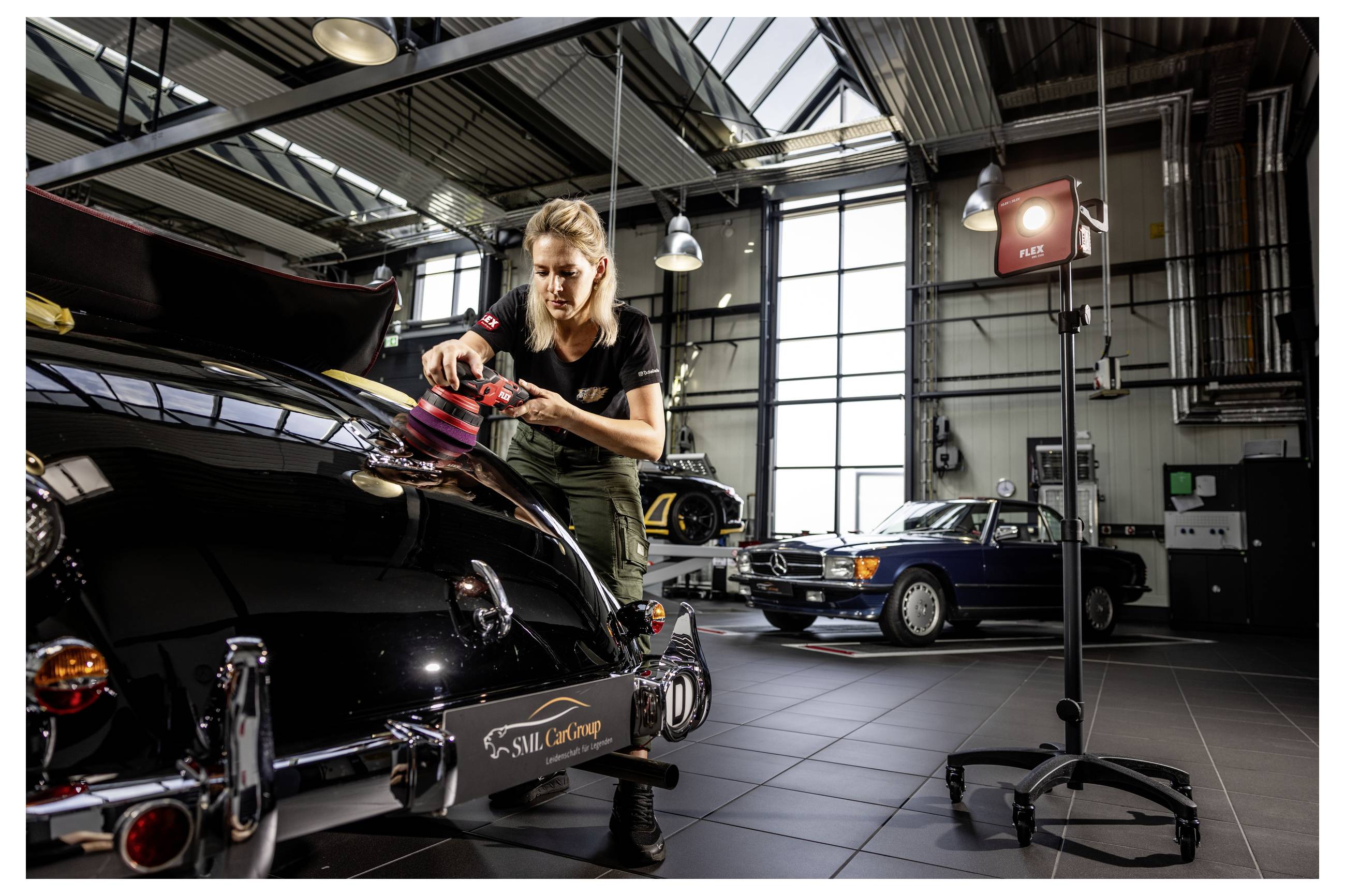 Ein Mechaniker poliert die Motorhaube eines schwarzen Klassickers in einer hellen Autowerkstatt, mit einem weiteren Oldtimer im Hintergrund.