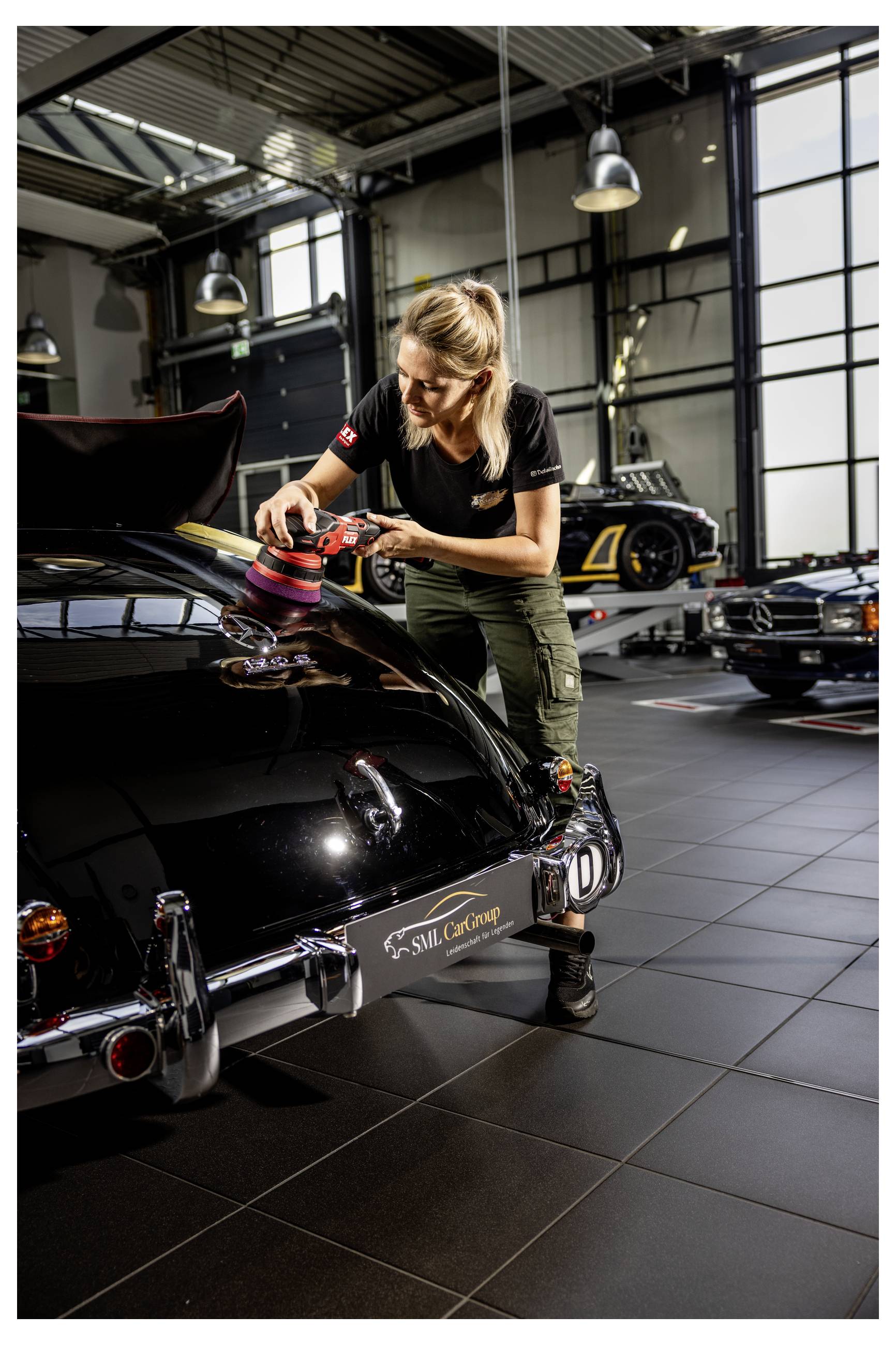 Frau poliert das Heck eines schwarzen Oldtimers in einer Werkstatt, mit anderen klassischen Autos und Werkstattleuchten im Hintergrund.
