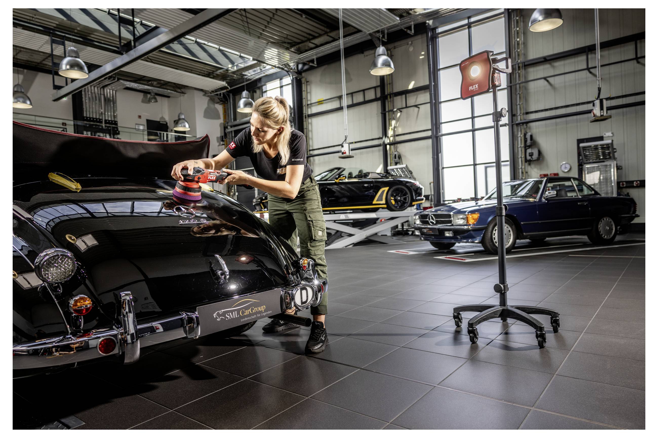 Frau poliert ein schwarzes Oldtimer-Auto mit einem Poliergerät in einer hell beleuchteten Autowerkstatt, mit anderen Vintage-Autos im Hintergrund.