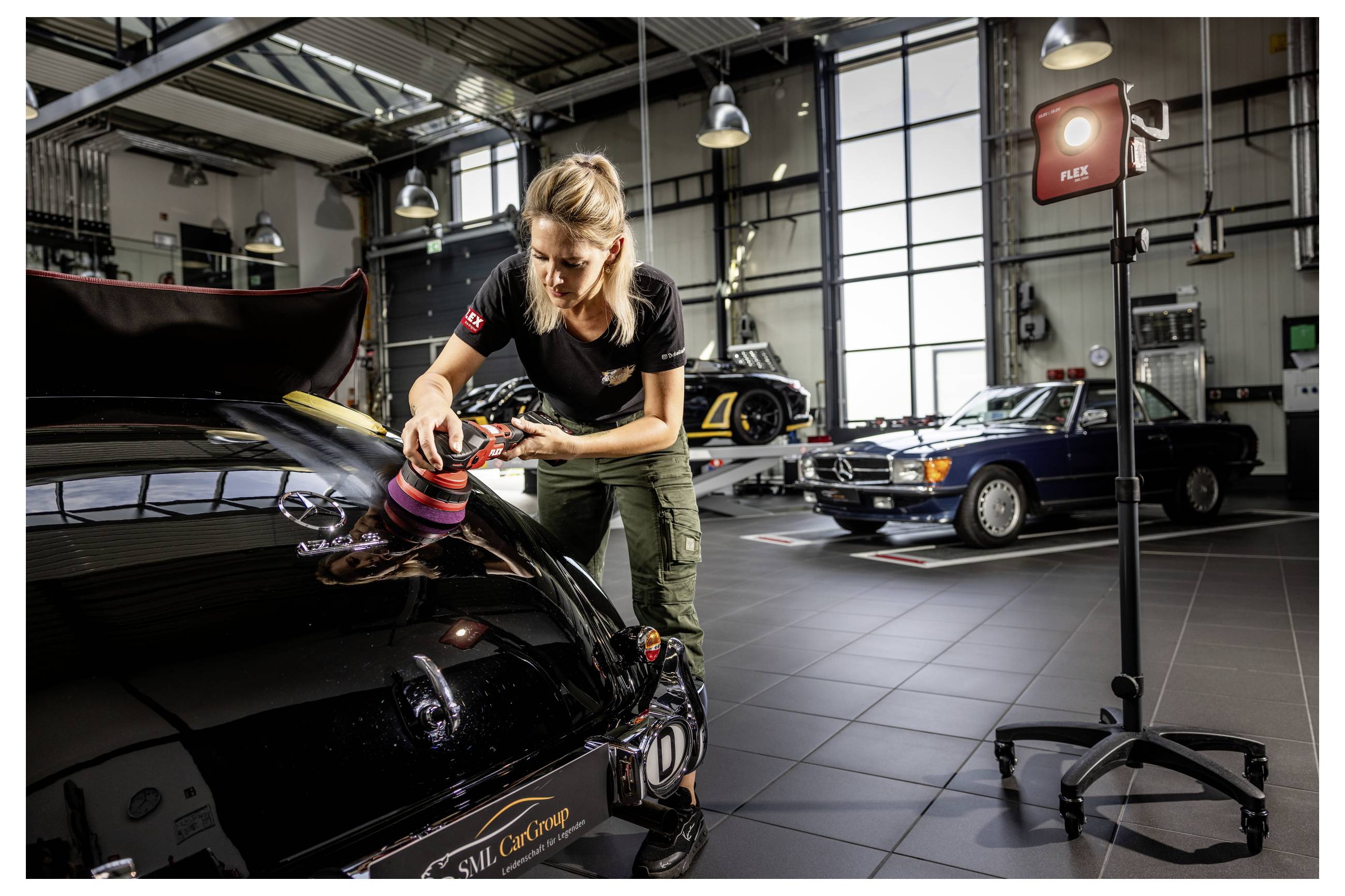 Frau poliert die Motorhaube eines schwarzen Autos in einer Garage-Werkstatt, während andere Klassiker im Hintergrund geparkt sind.