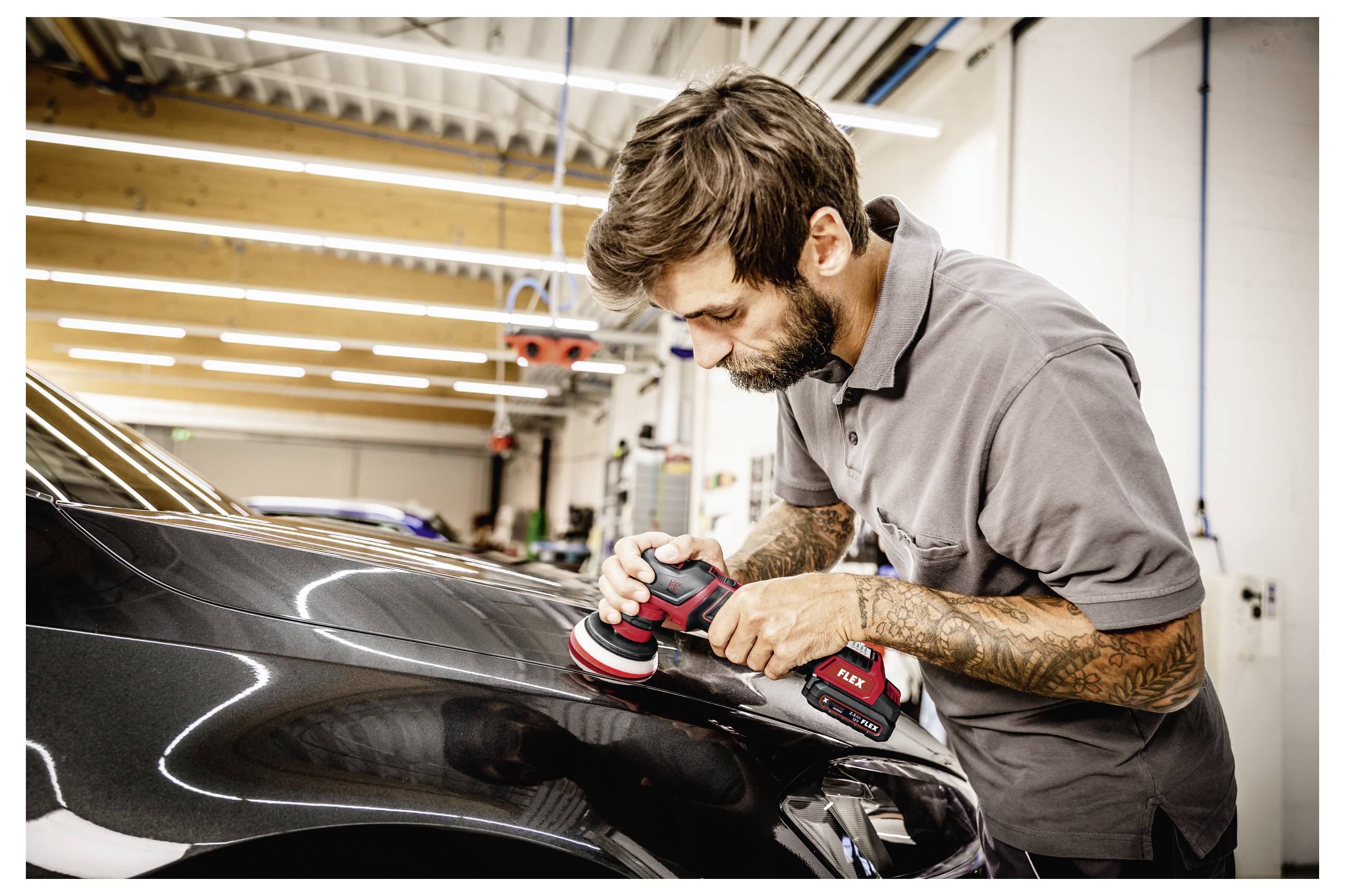 Ein Mann poliert die Motorhaube eines schwarzen Autos mit einem tragbaren Poliergerät in einer Autowerkstatt.