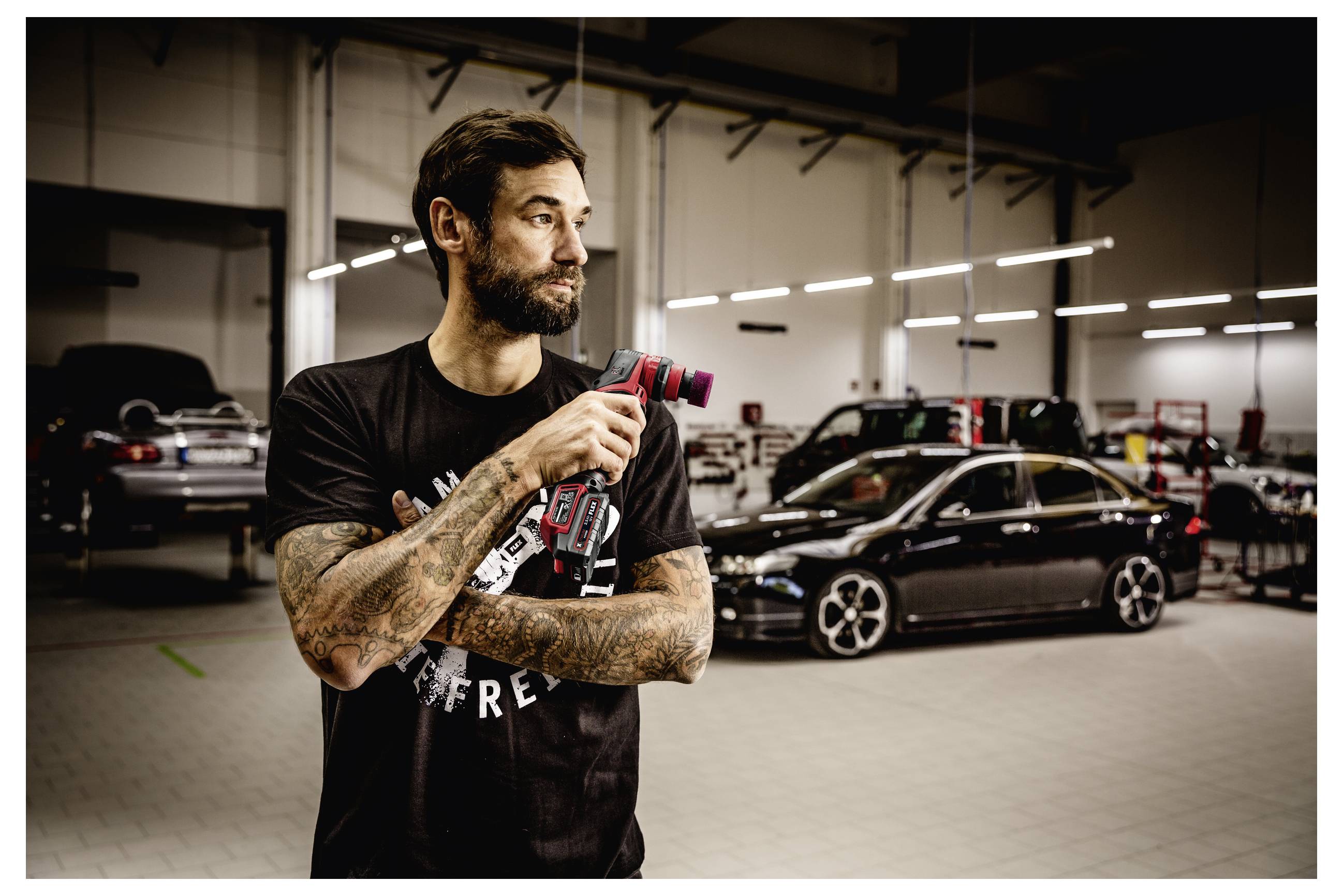 Tätowierter Mann, der ein rotes Elektrowerkzeug in einer Autowerkstatt hält, mit mehreren schwarzen Autos, die hinter ihm geparkt sind.